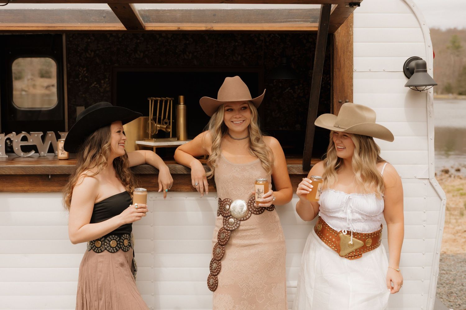Three friends in brown dresses and cowboy hats gather together enjoying drinks outside a vintage trailer.