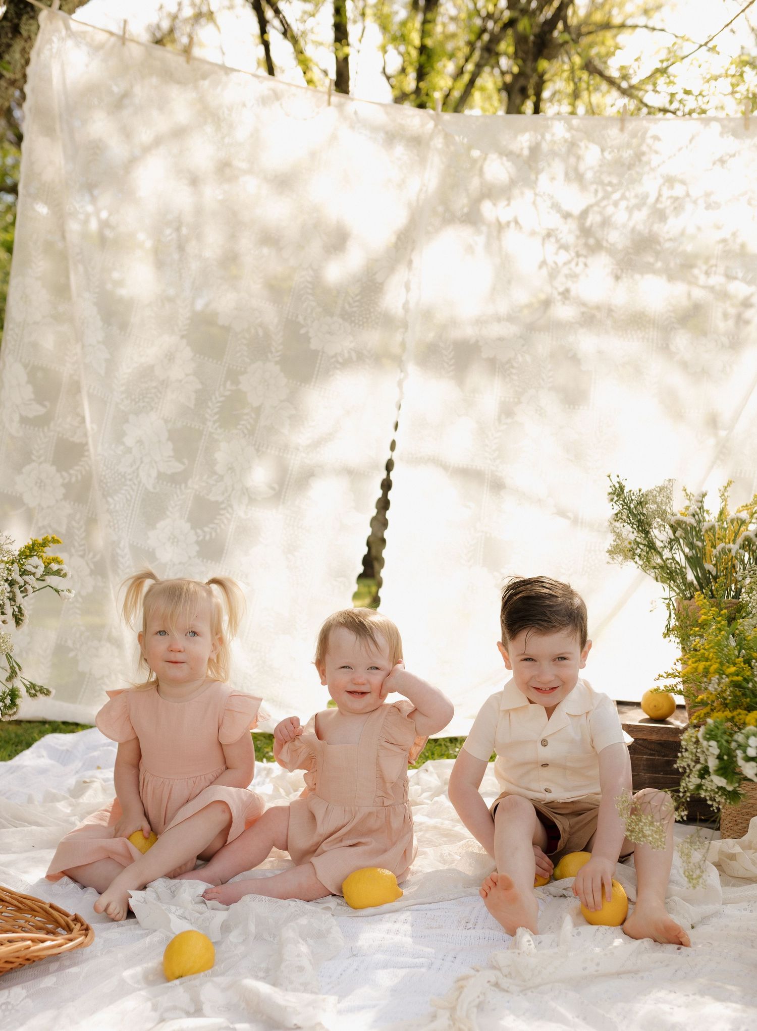 Three young children sit on a white blanket outdoors with lemons, posed in front of a sheer floral backdrop.