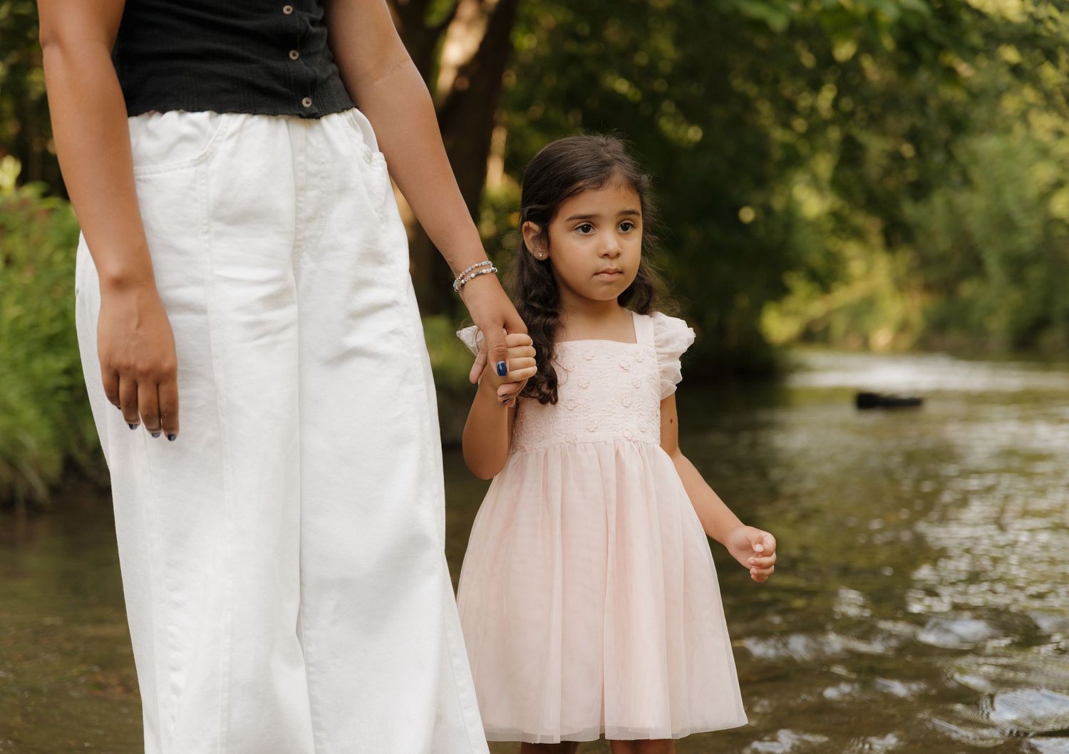 A young child in a pink dress holds hands with someone while standing by a river.