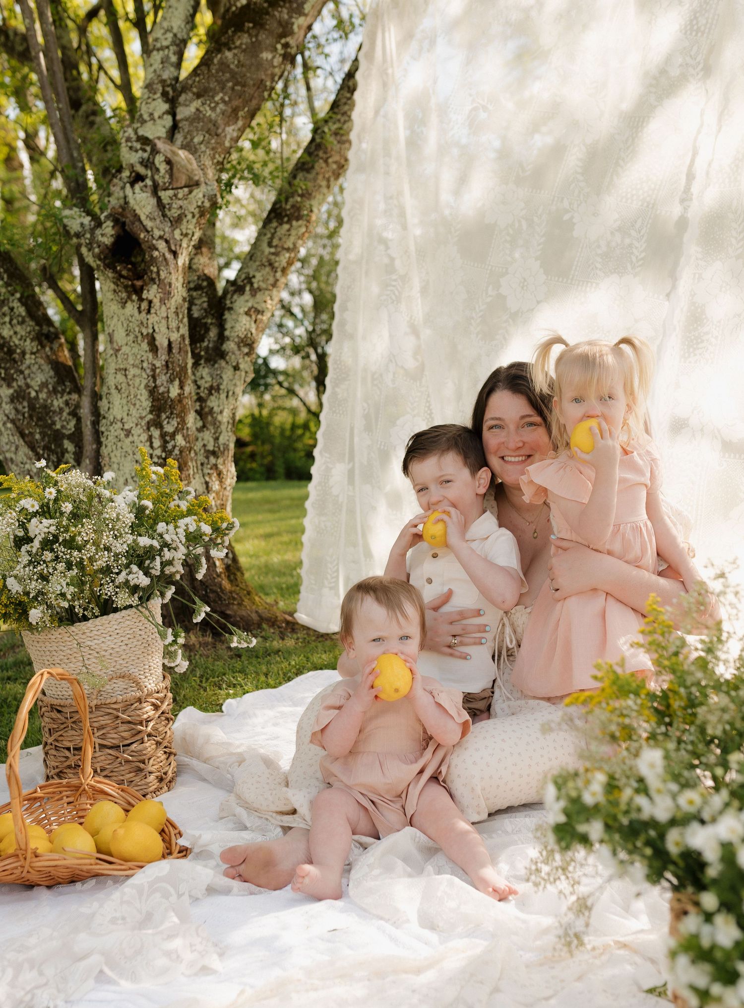 A mother sits outdoors on a white blanket with her three young children, all holding lemons near a floral backdrop.