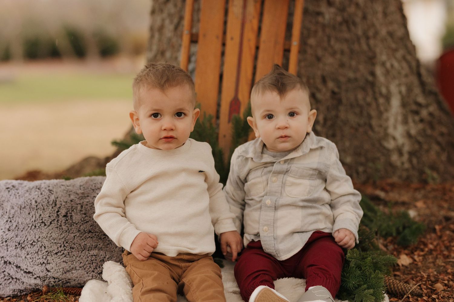 Two babies wearing matching cream colored outfits sit together on a plush gray chair.