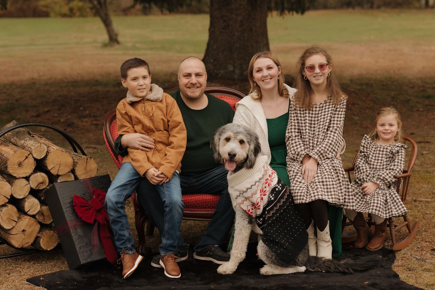 A family sits on a rustic bench with their golden retriever dog next to stacked firewood in an autumn setting.