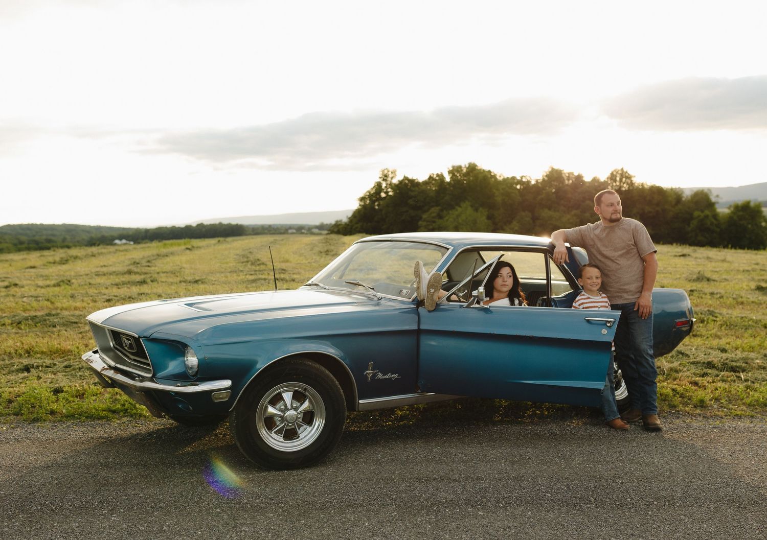 Family posing with a classic blue Ford Mustang in a scenic open field at sunset.
