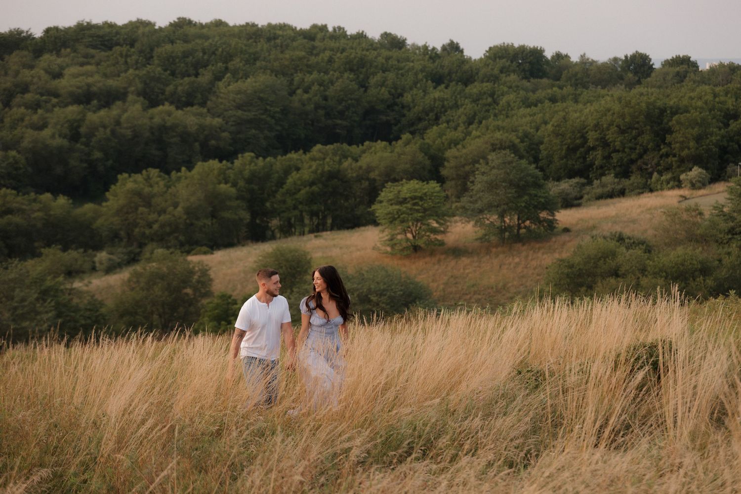 Couple walking through tall golden grass with rolling hills in background.