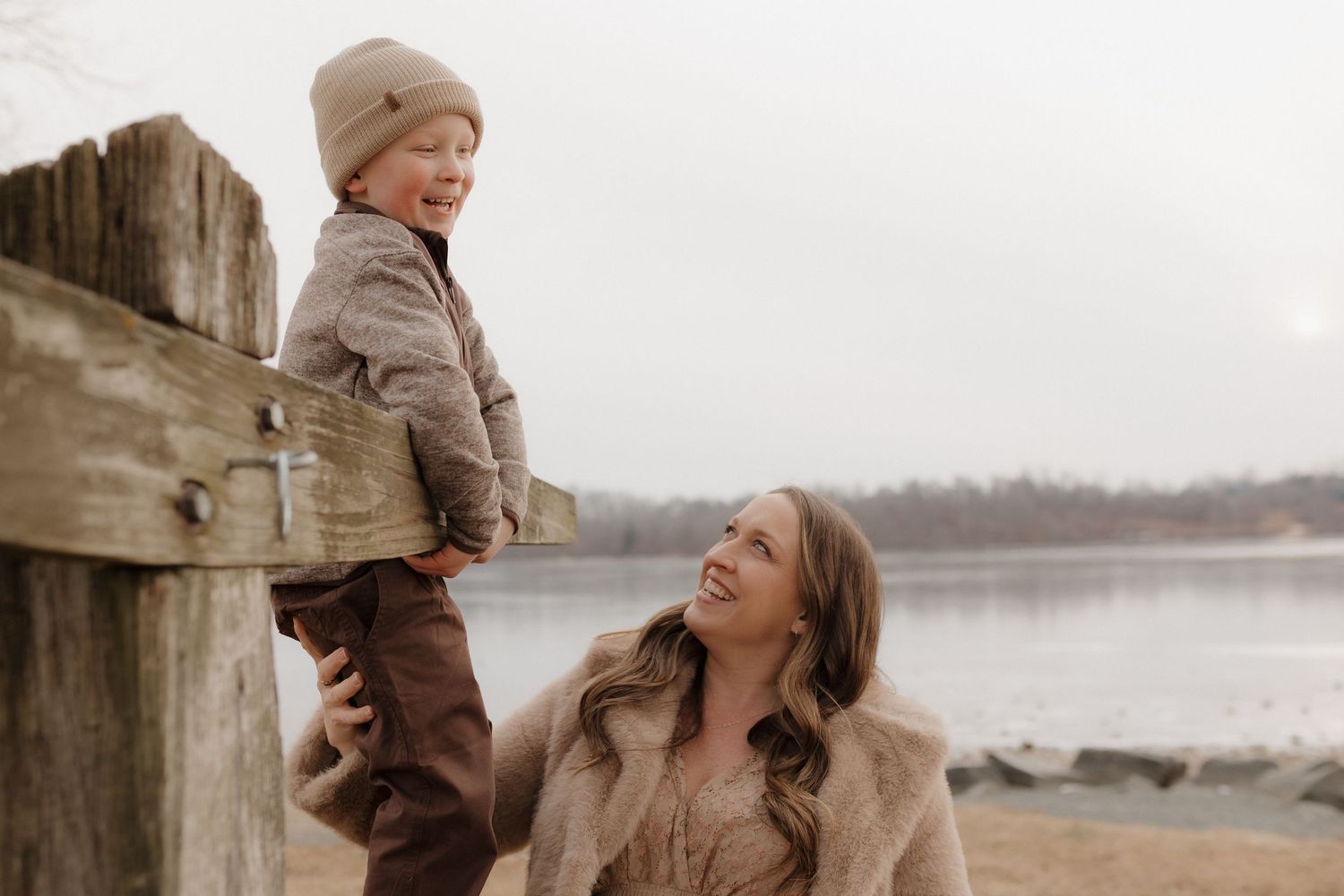A smiling mother and young child in warm winter clothing enjoy a playful moment by a lakeside wooden dock.