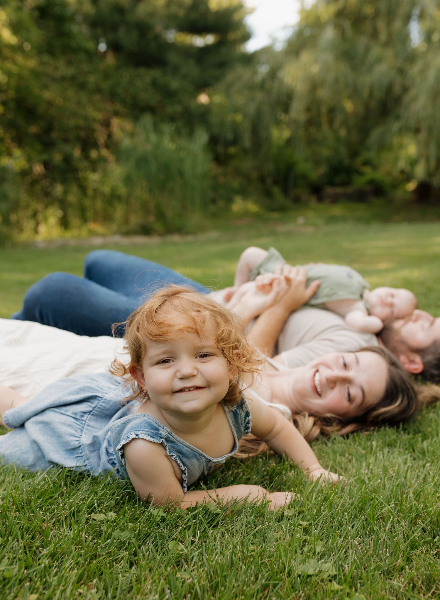 A happy toddler girl with curly red hair lies on green grass while her smiling family relaxes together outdoors.