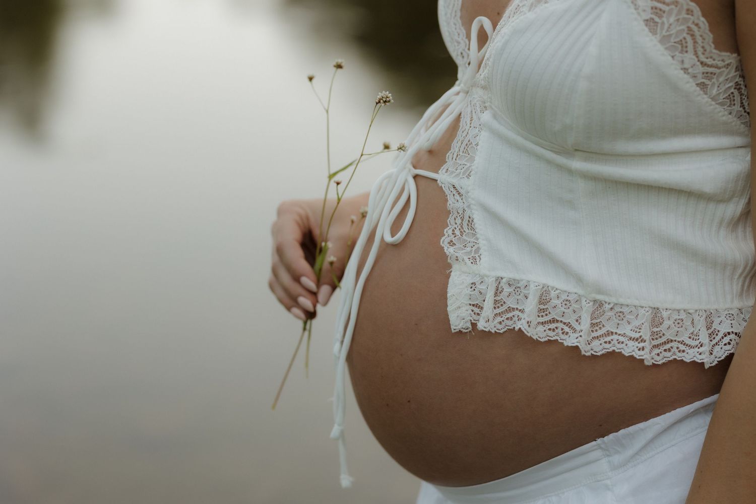 Close-up of a pregnant belly in white lace maternity wear with delicate dried flowers.