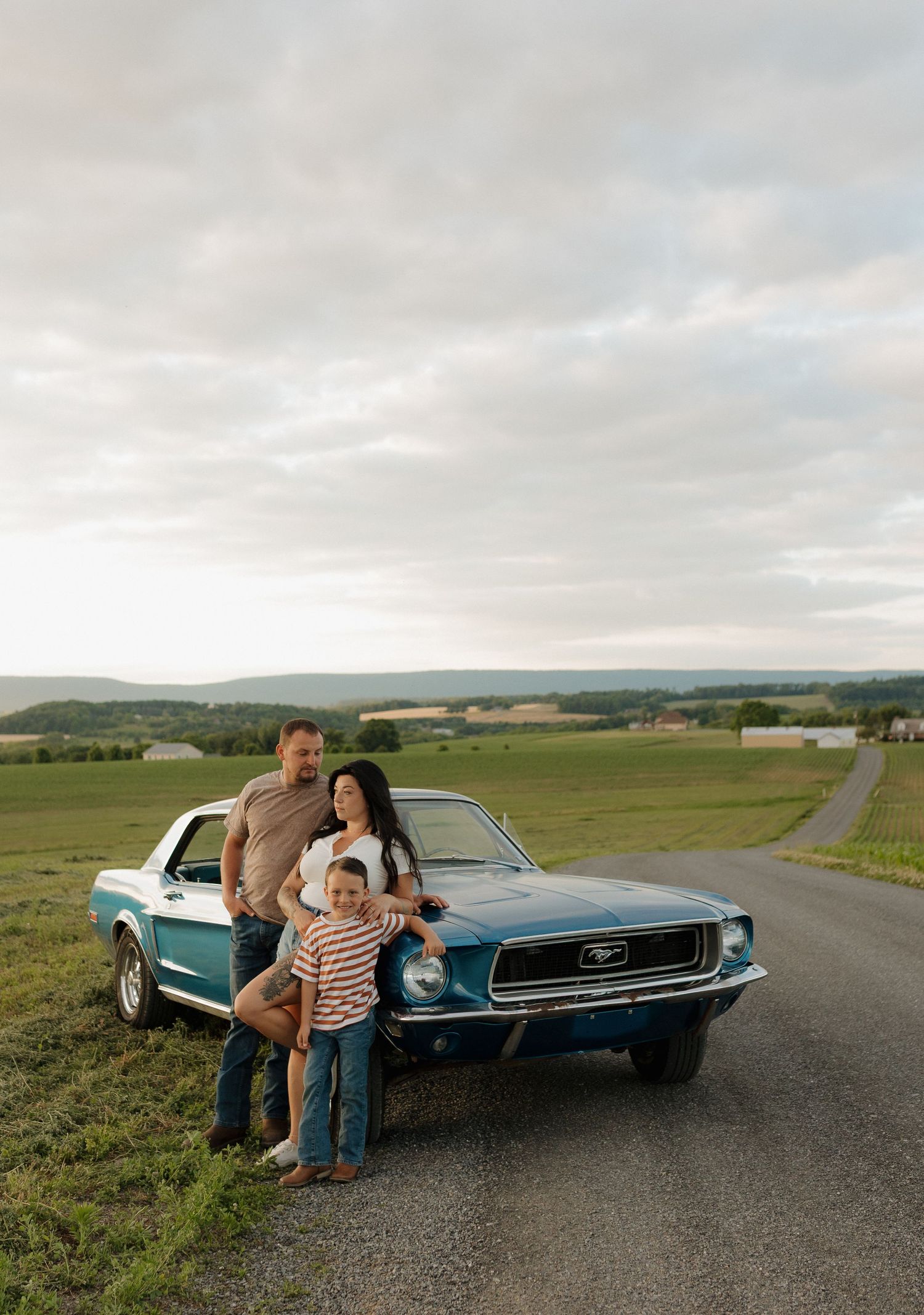 Family posing with a classic blue Ford Mustang on a rural road surrounded by green fields under a cloudy sky.