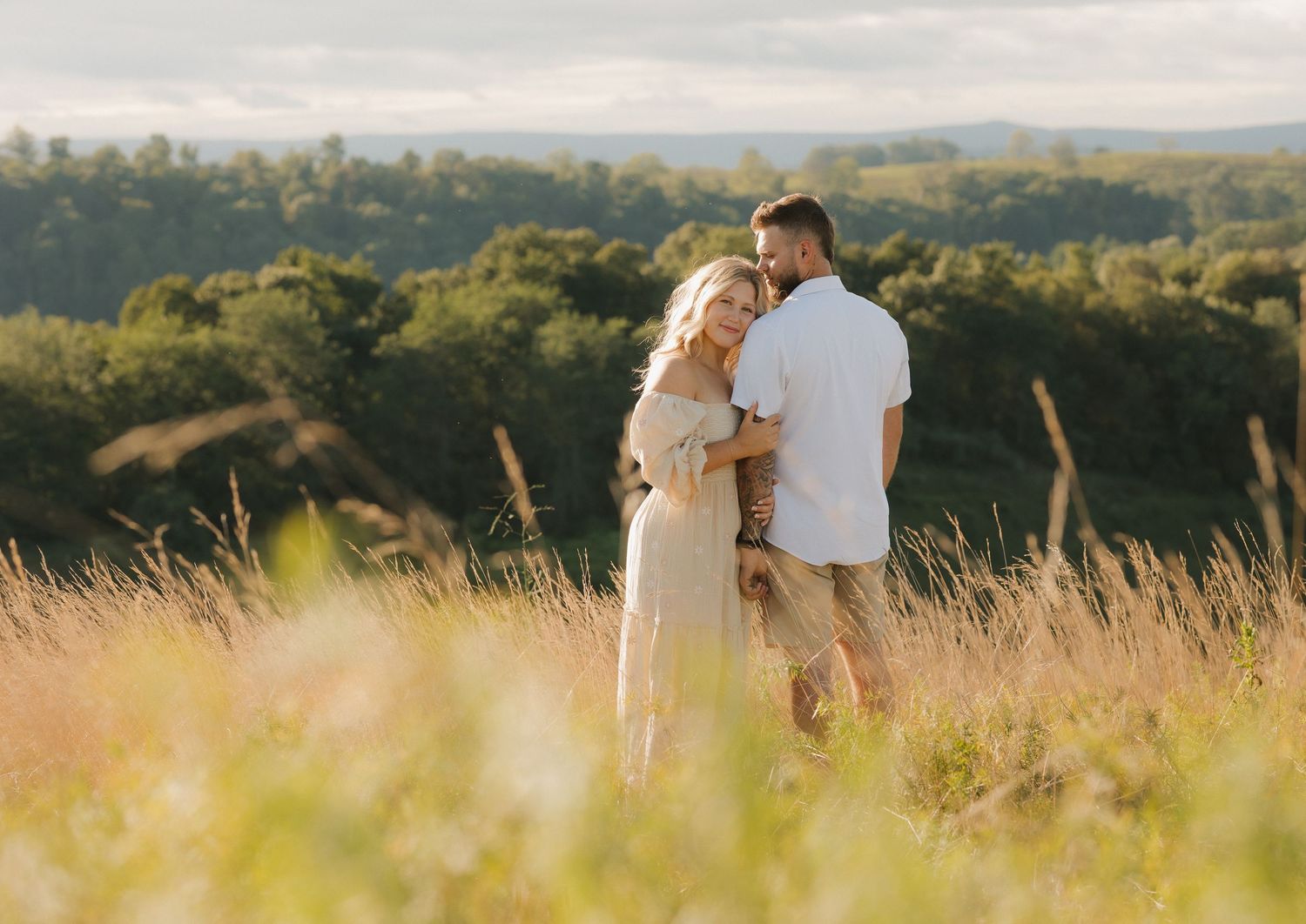 Couple embraces in golden sunlit meadow during sunset with rolling hills in background.