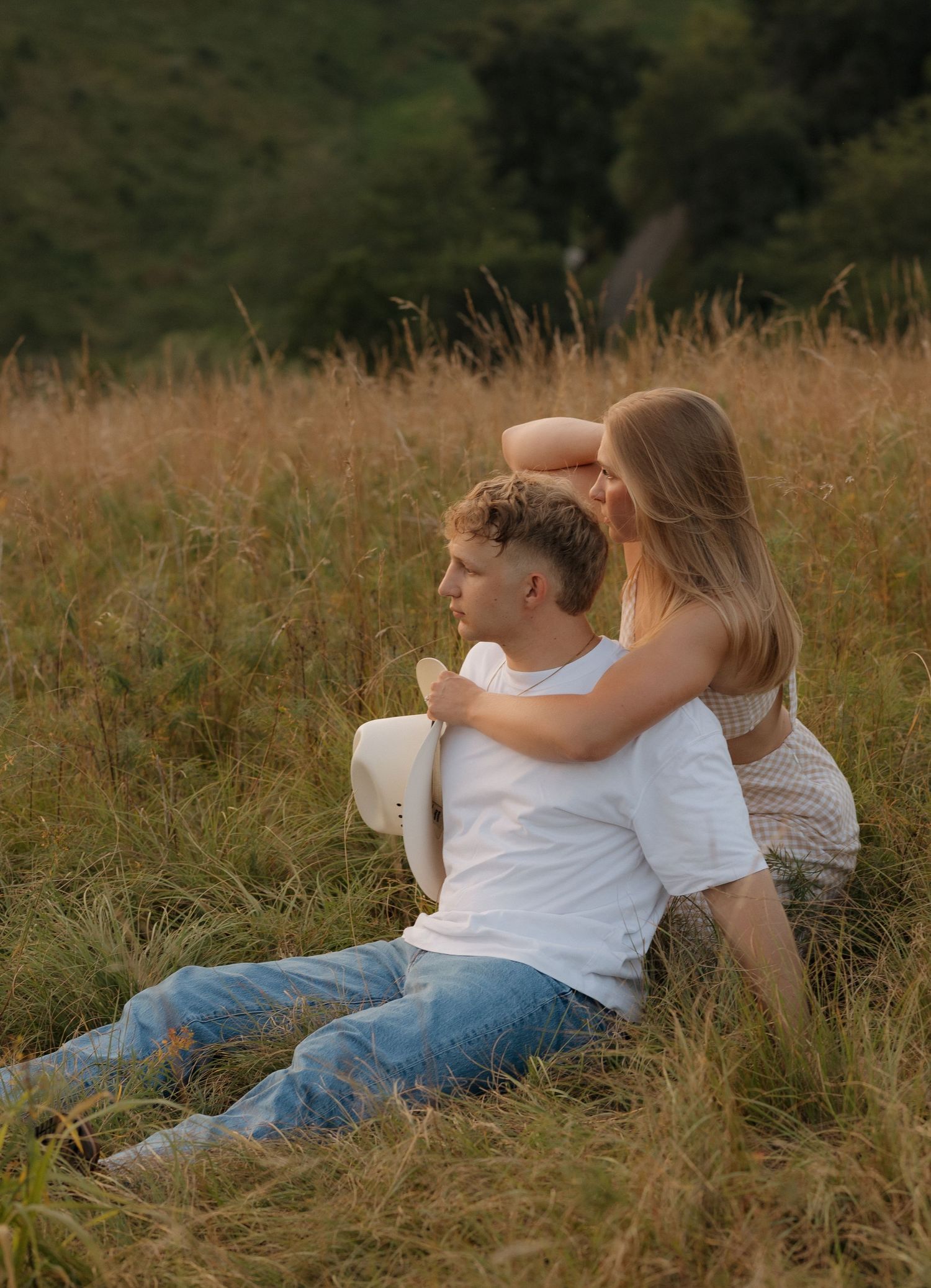 A couple sits together in tall grass on a hillside at sunset.