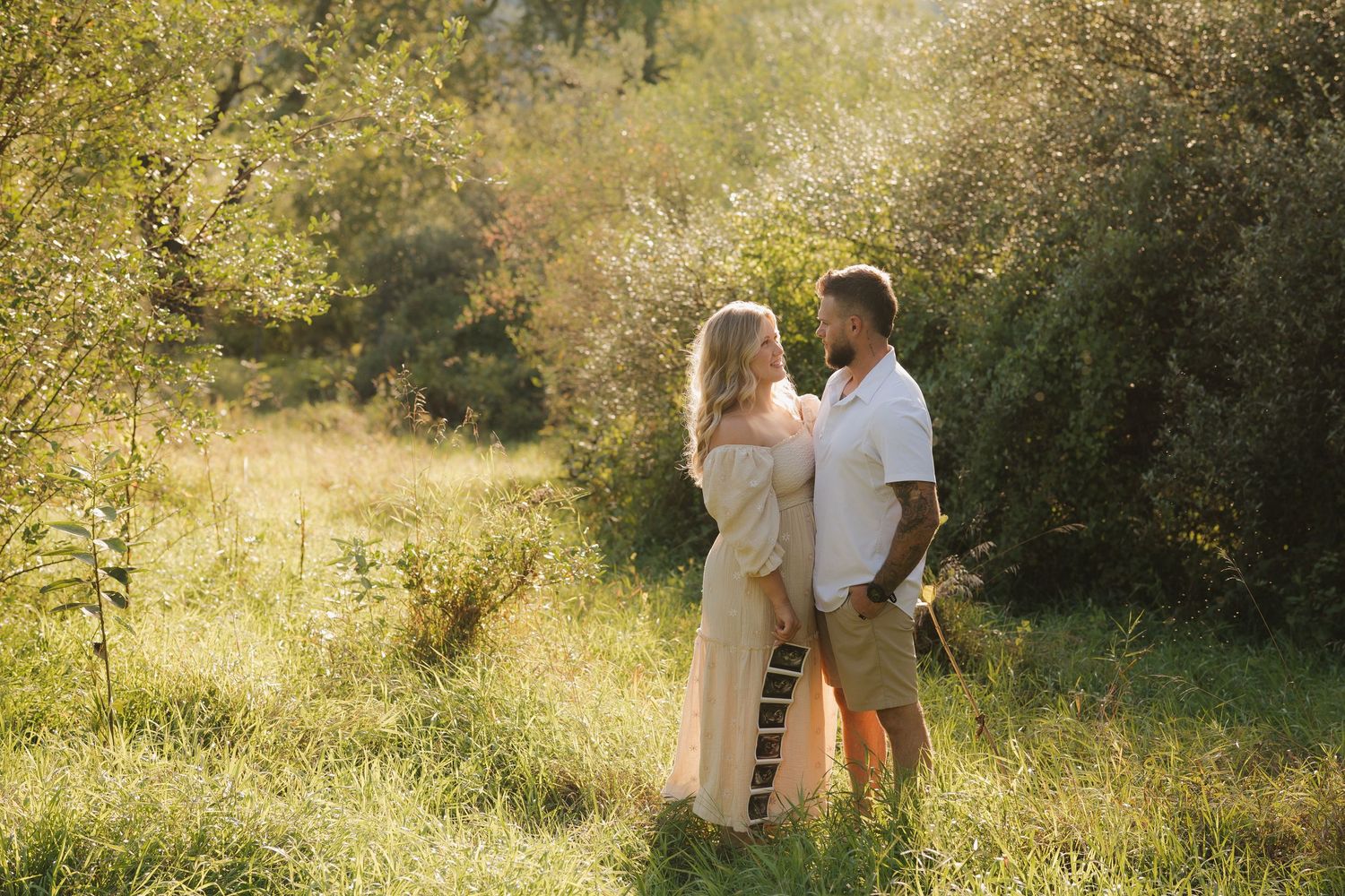 A couple shares a tender moment in a sunlit meadow surrounded by lush greenery and golden afternoon light.