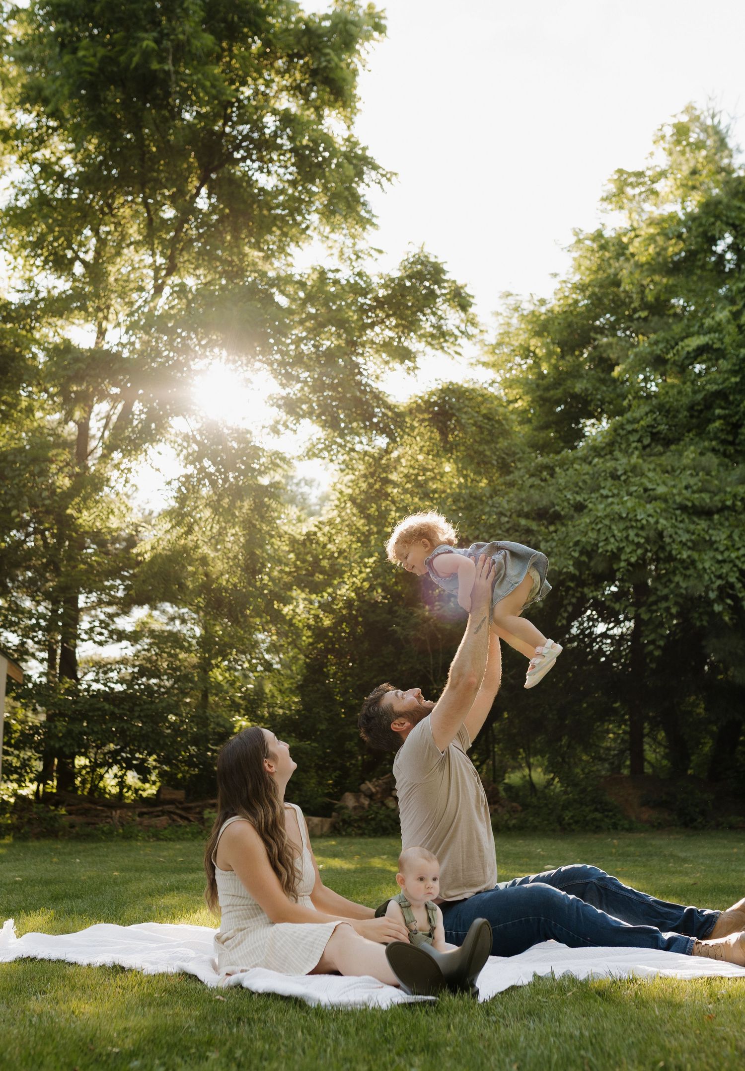 Family enjoying golden hour outdoors on a blanket, father lifting baby in the air while mother sits smiling nearby.