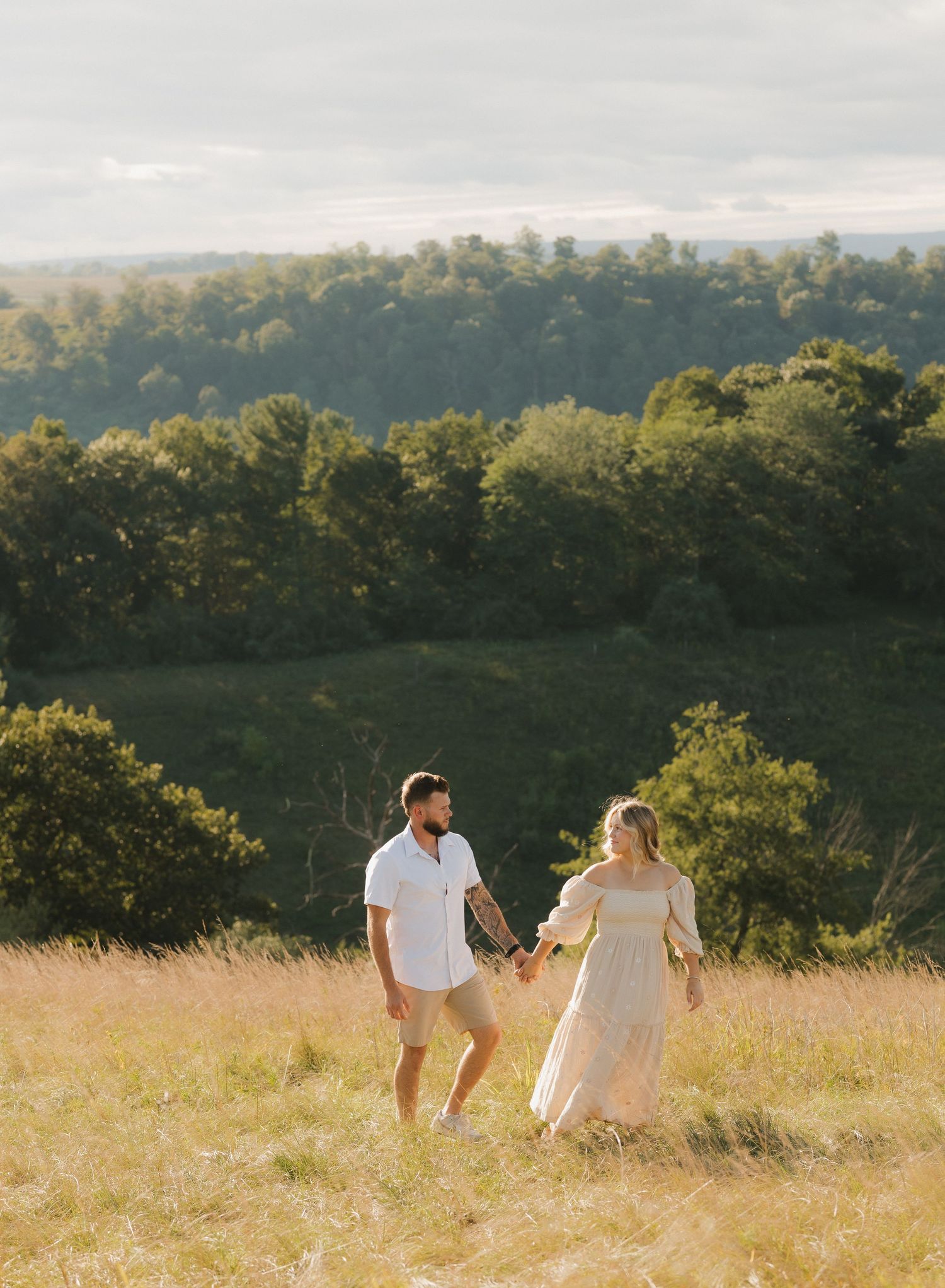 A couple walks hand in hand through a sunlit meadow with rolling hills and forest in the background during golden hour.