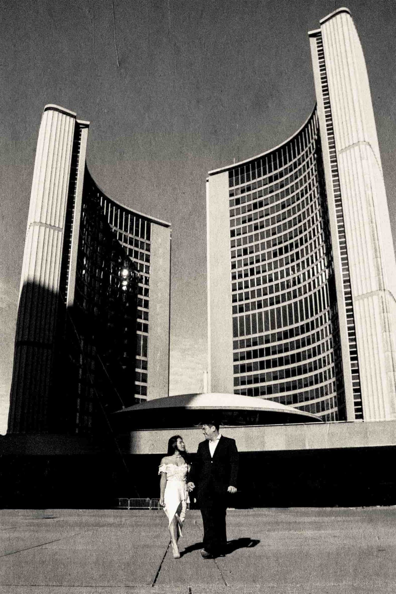 Black and white photo of couple walking towards camera with Toronto City Hall behind them.
