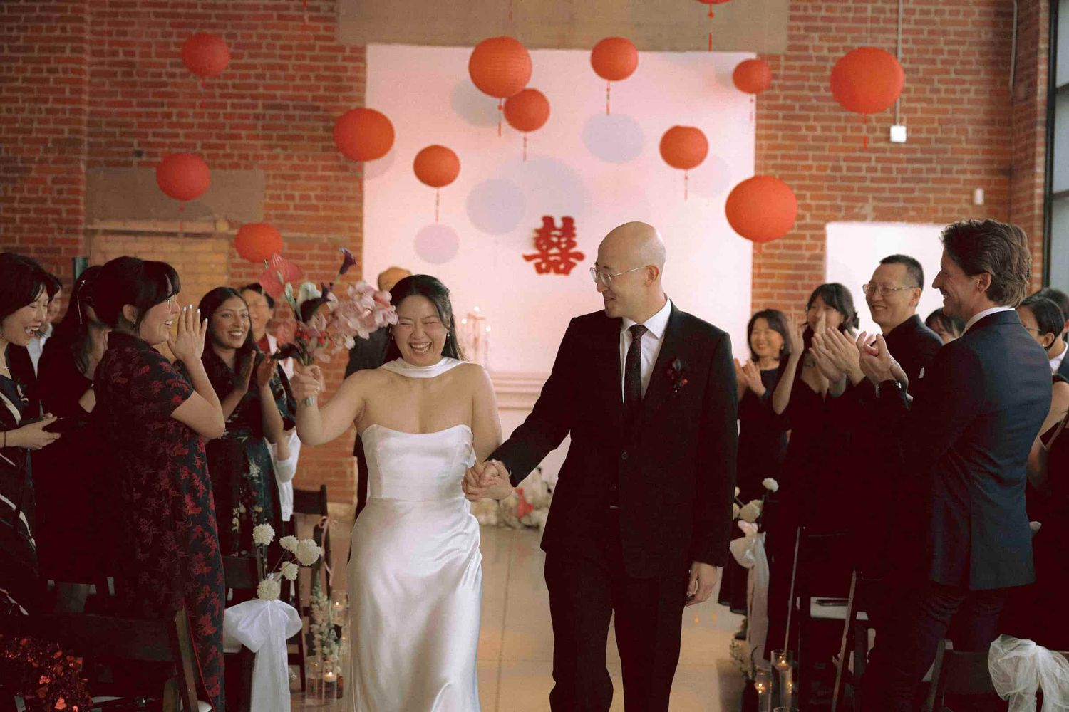 Newly married couple walking down the aisle beneath red lantern decorations after their ceremony.