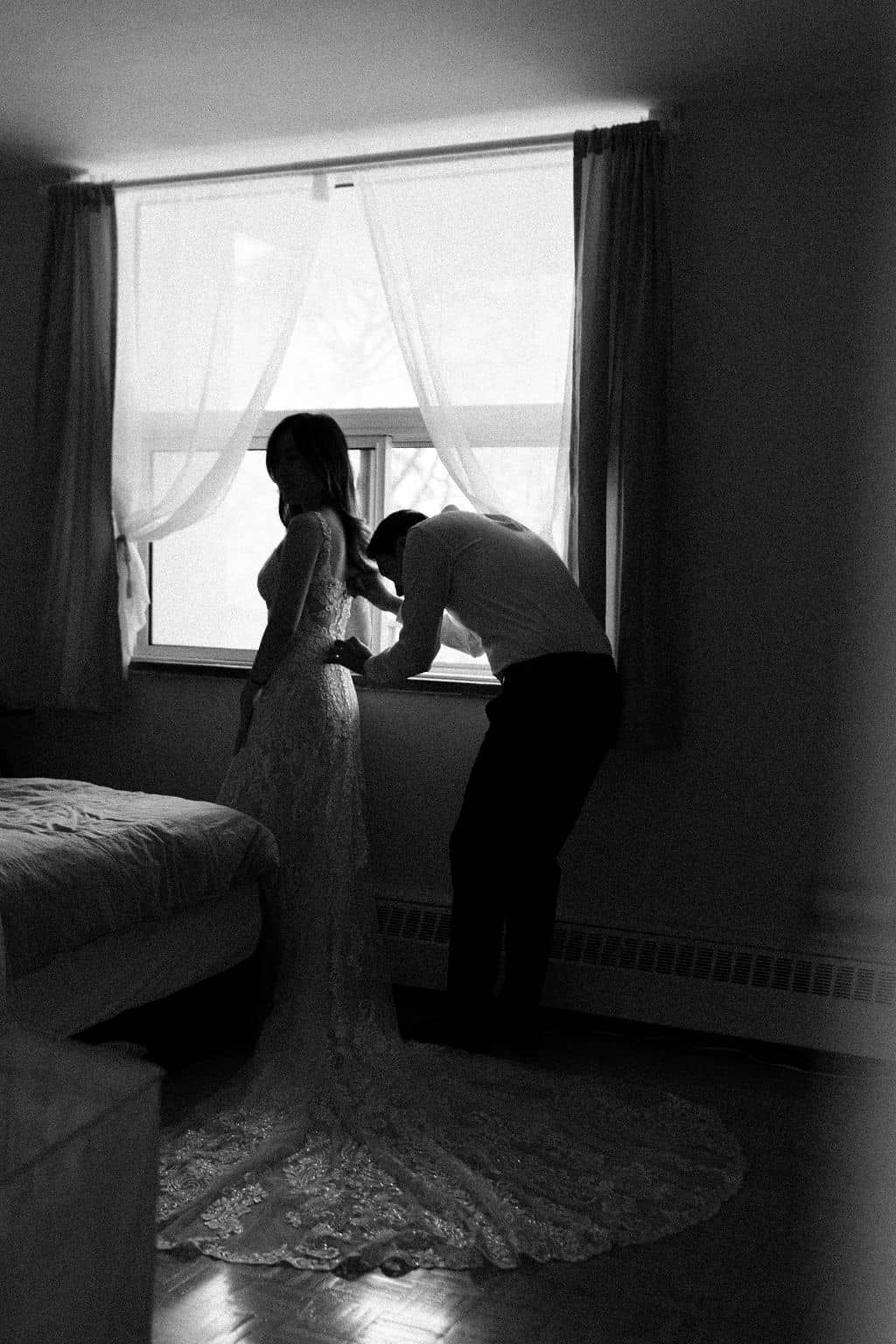 groom helping bride with her dress as they get ready together at home before their elopement at Scarborough Bluffs