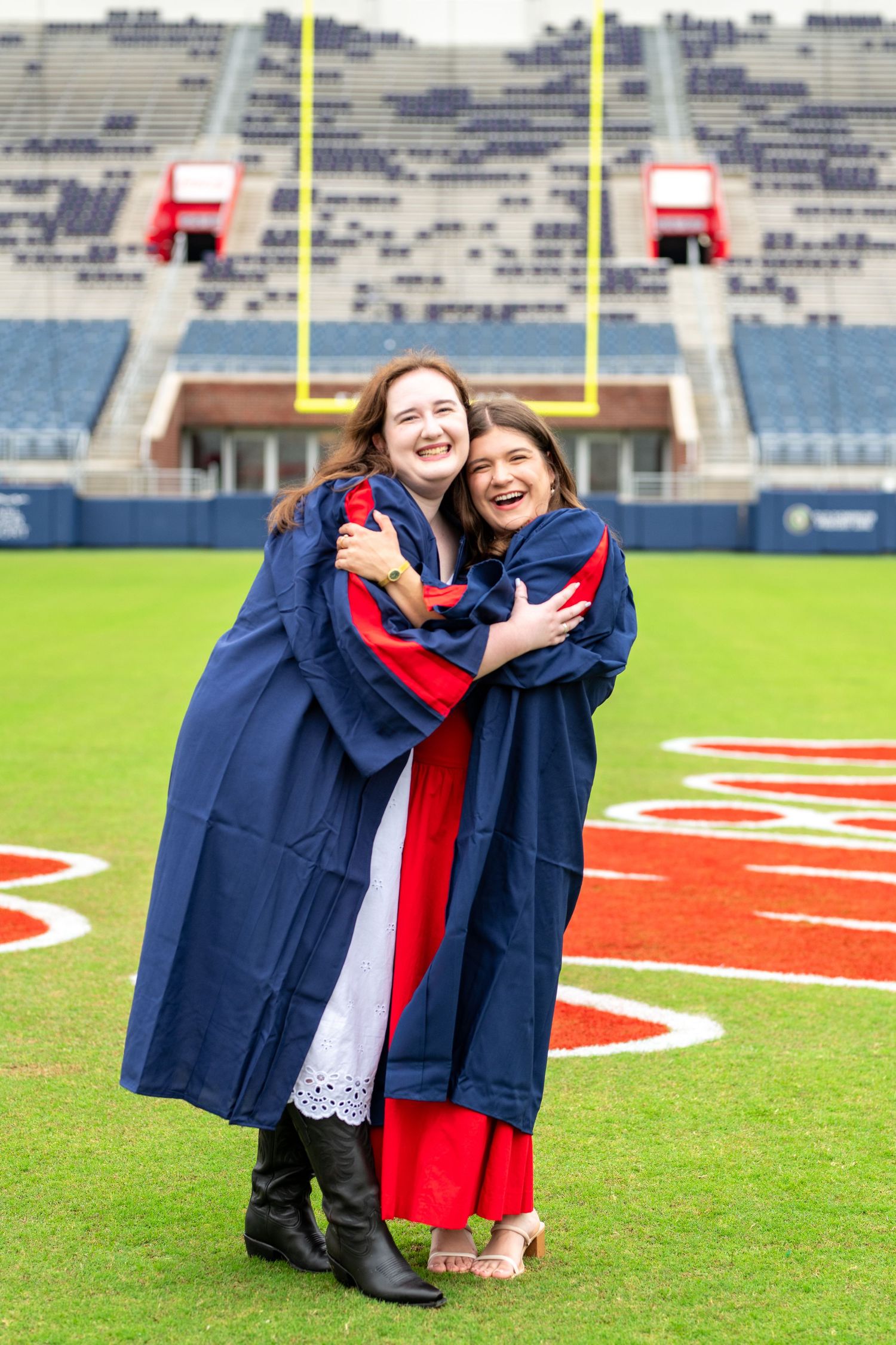 Two graduates in navy and red regalia embrace on football field with stadium in background.
