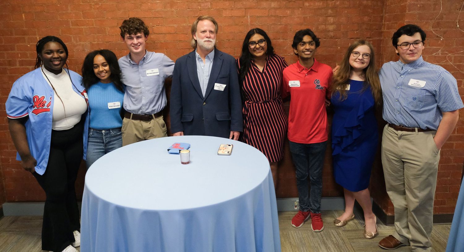 A diverse group of eight students and a faculty member pose together around a tall round table at a formal academic event.
