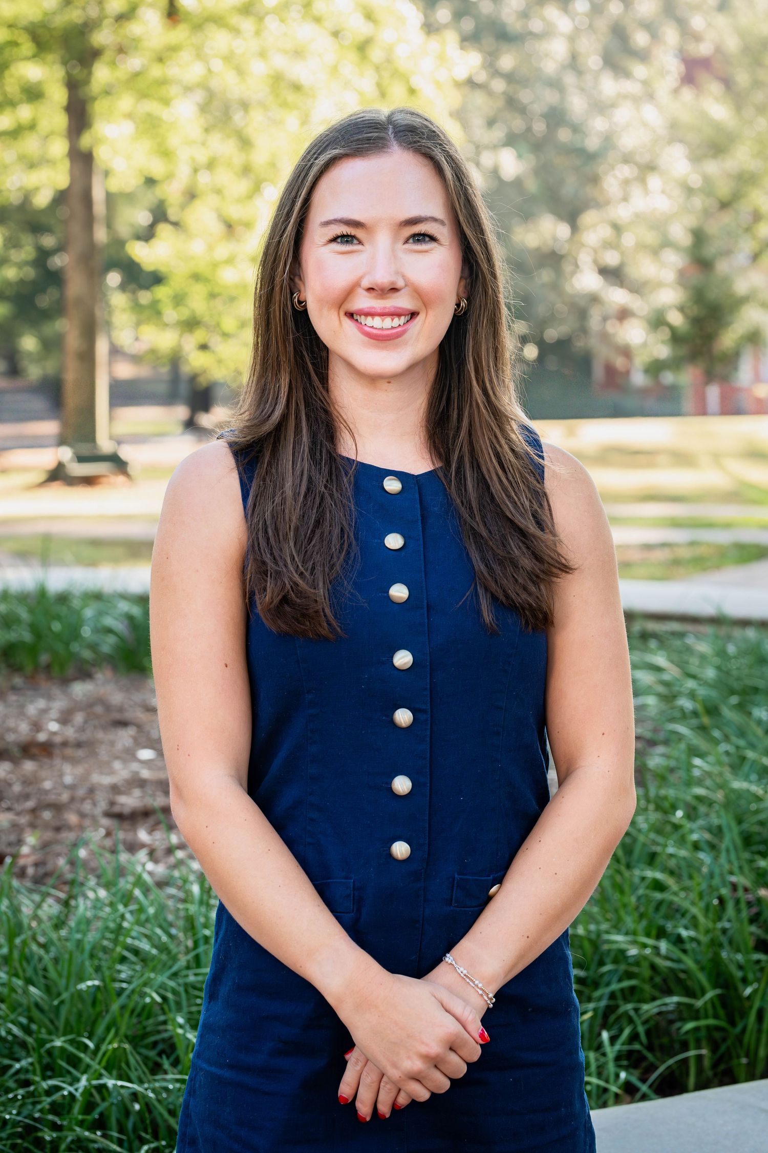 Smiling young woman in navy blue sleeveless dress standing outdoors on a sunny day near green trees.