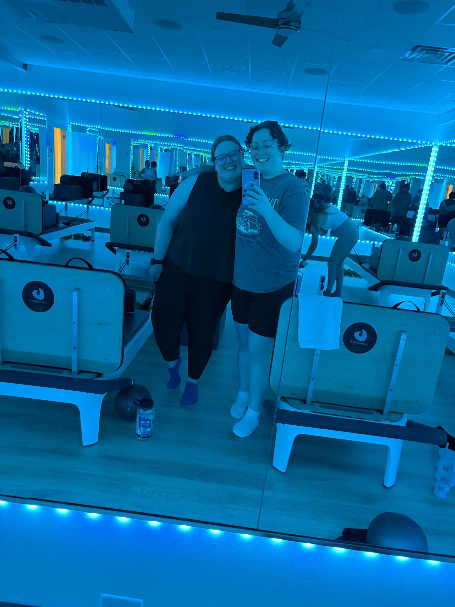 Two women pose for a mirror selfie in a blue neon-lit fitness studio with rowing machines.