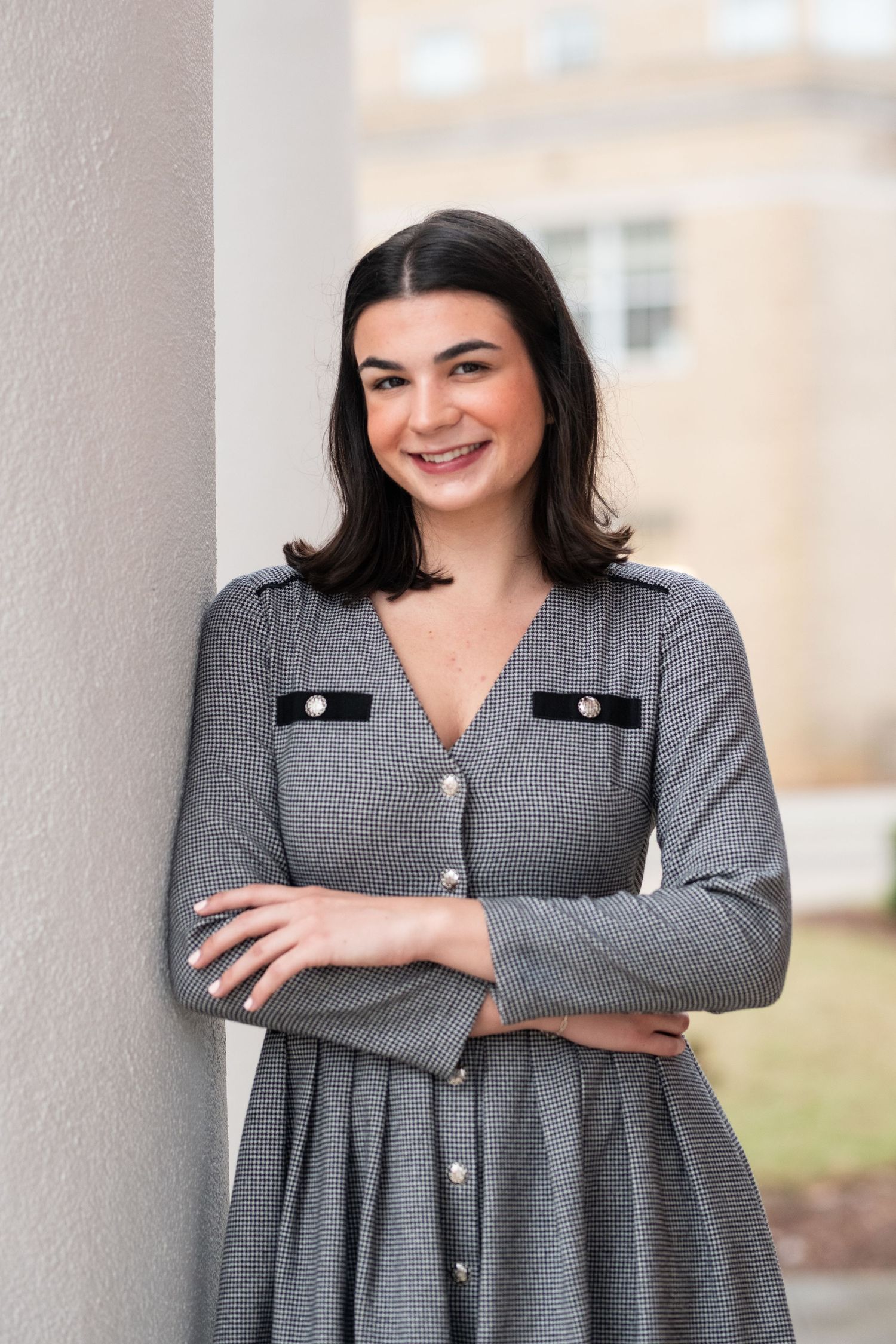 A person wearing a gray pleated dress with decorative buttons leans against a white pillar outdoors.