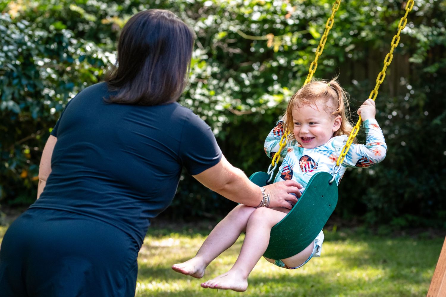 A mother pushes her happy toddler girl on a green garden swing outdoors.