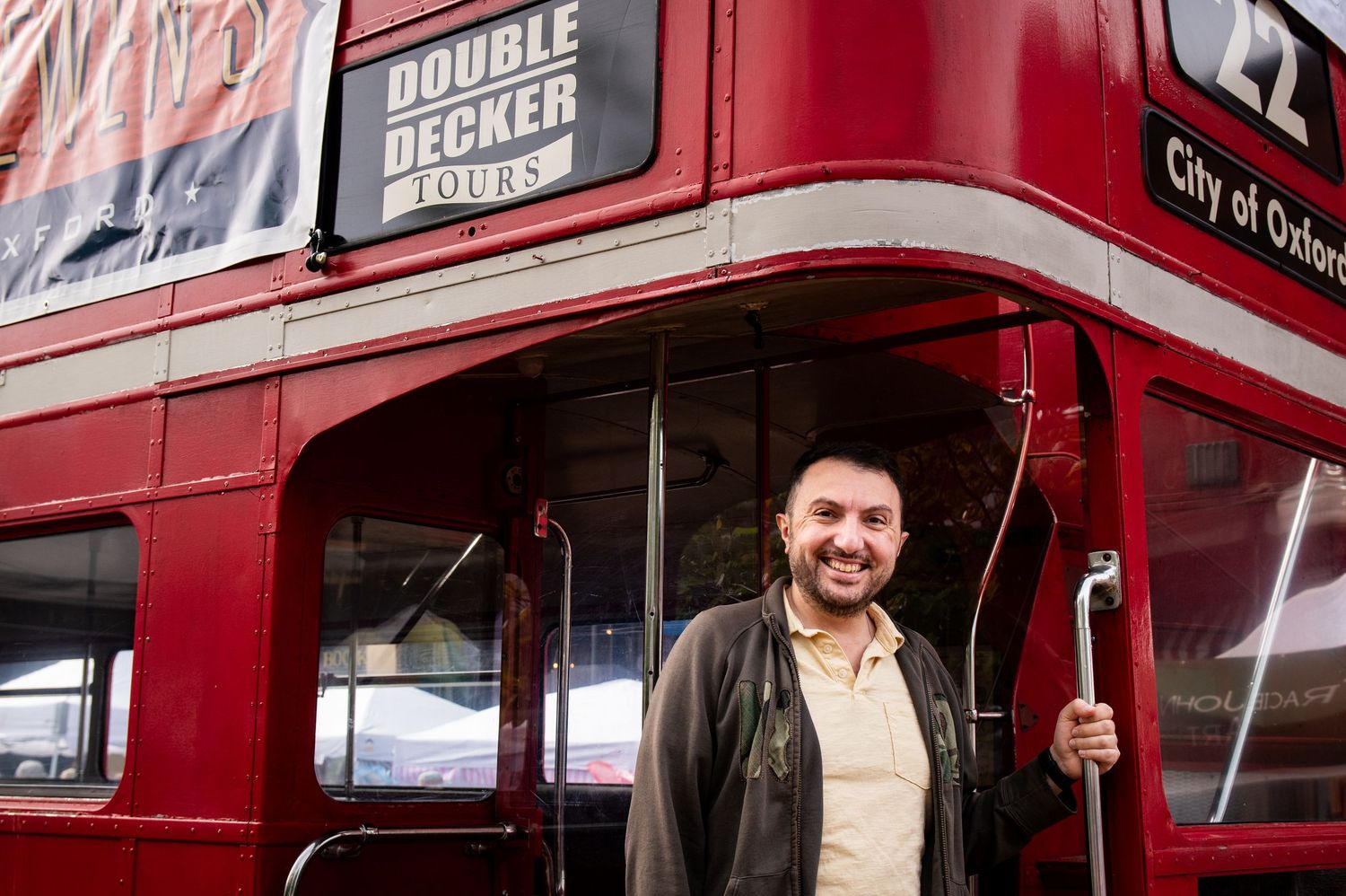 Man smiling on a red Double Decker Tours bus in City of Oshkosh, holding a pole at the entrance.