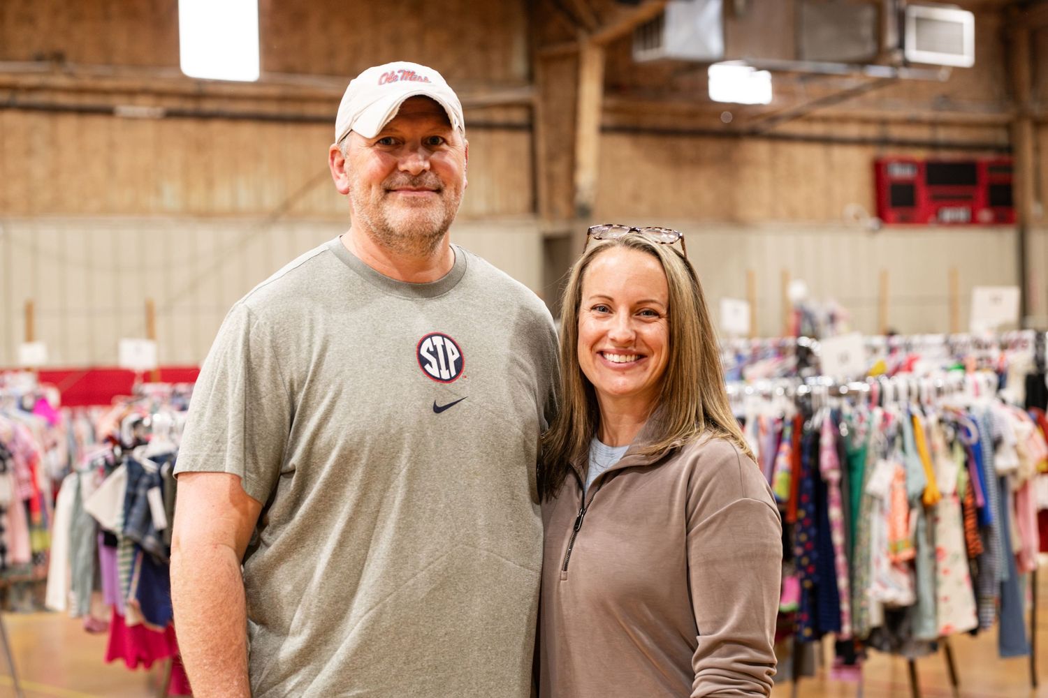 A smiling couple poses together inside a large indoor consignment sale with colorful clothing racks in the background.