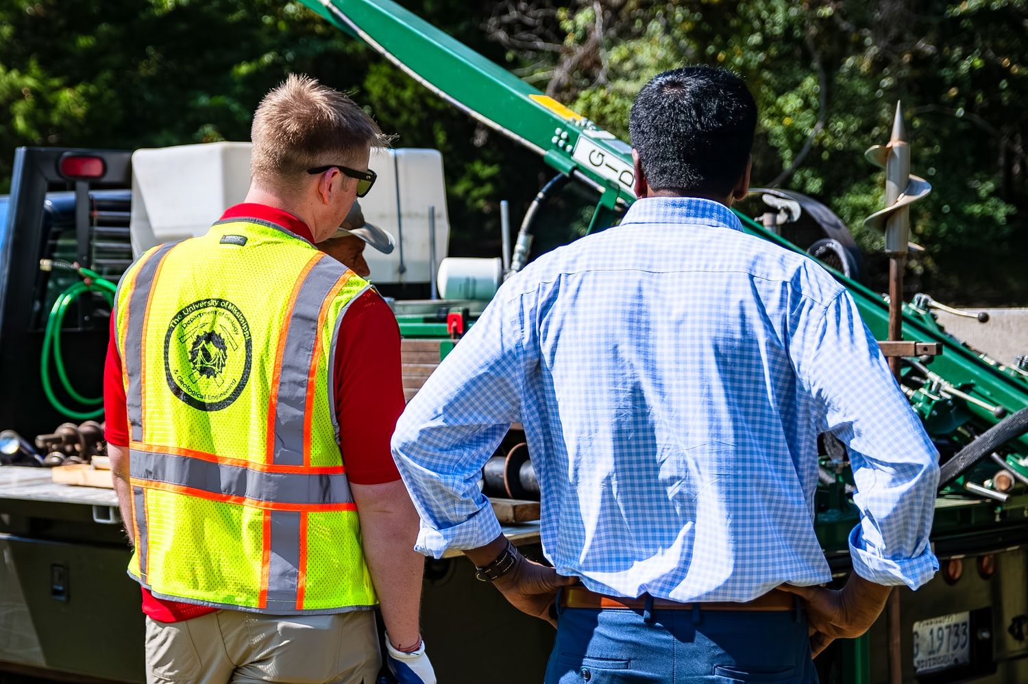 Two workers examining green construction equipment outdoors, one wearing a yellow safety vest.