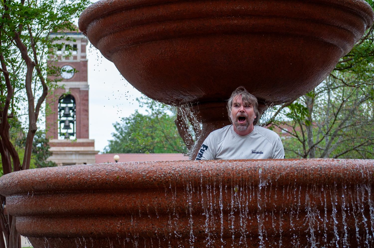 A person peers from behind a large terracotta fountain with cascading water in a park setting.