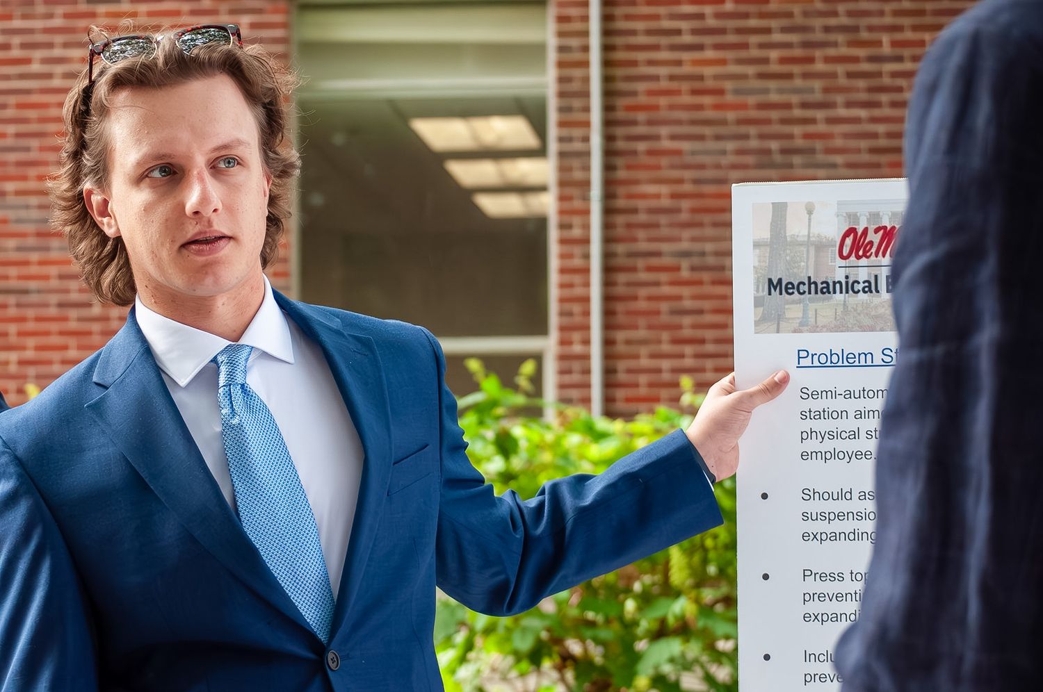 Man in blue suit pointing at a sign outside a university mechanical engineering building.