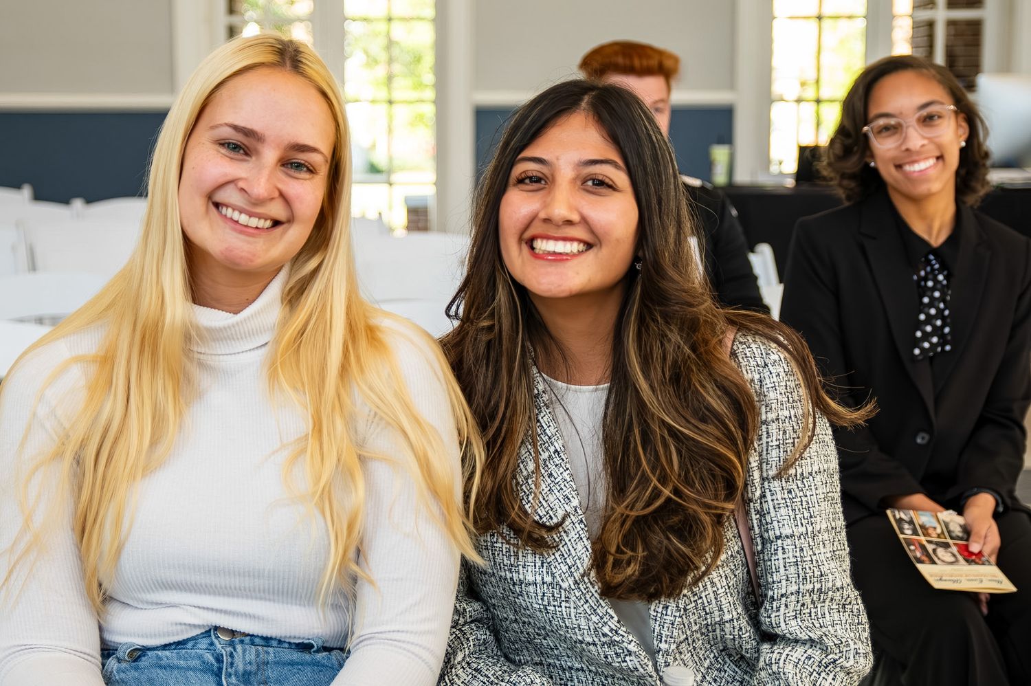 Three smiling young women sitting together at an indoor event, dressed in smart casual attire.