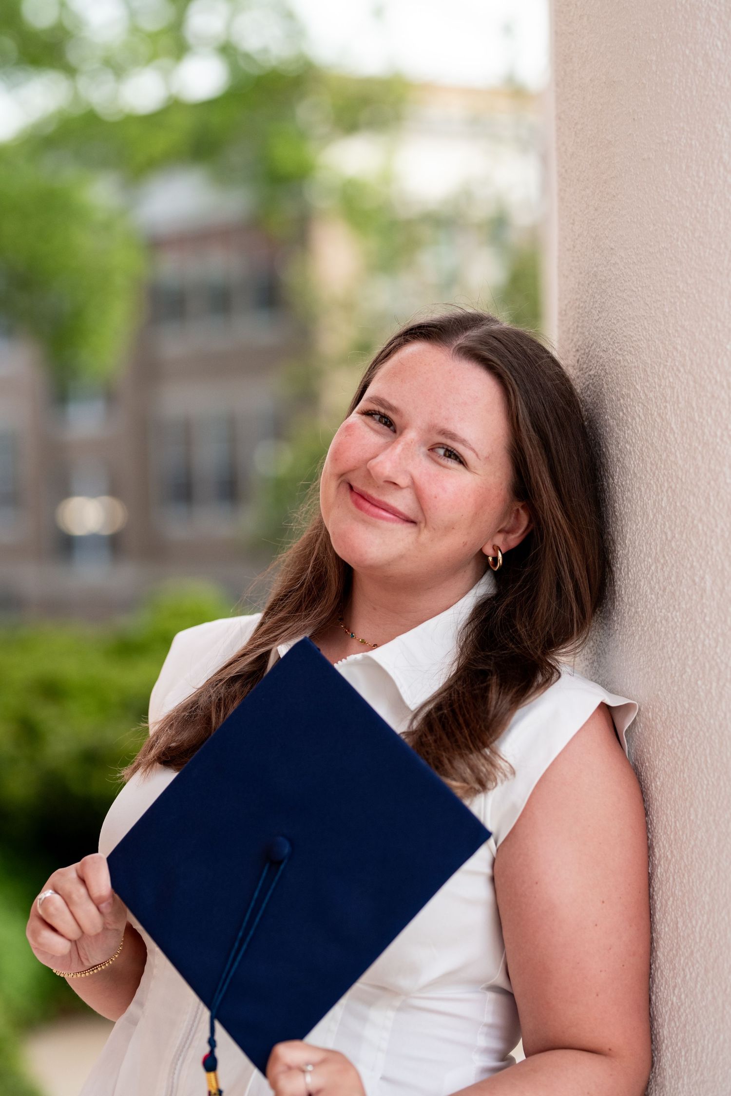 Smiling woman in white dress holding navy blue graduation cap leans against wall outdoors on college campus.