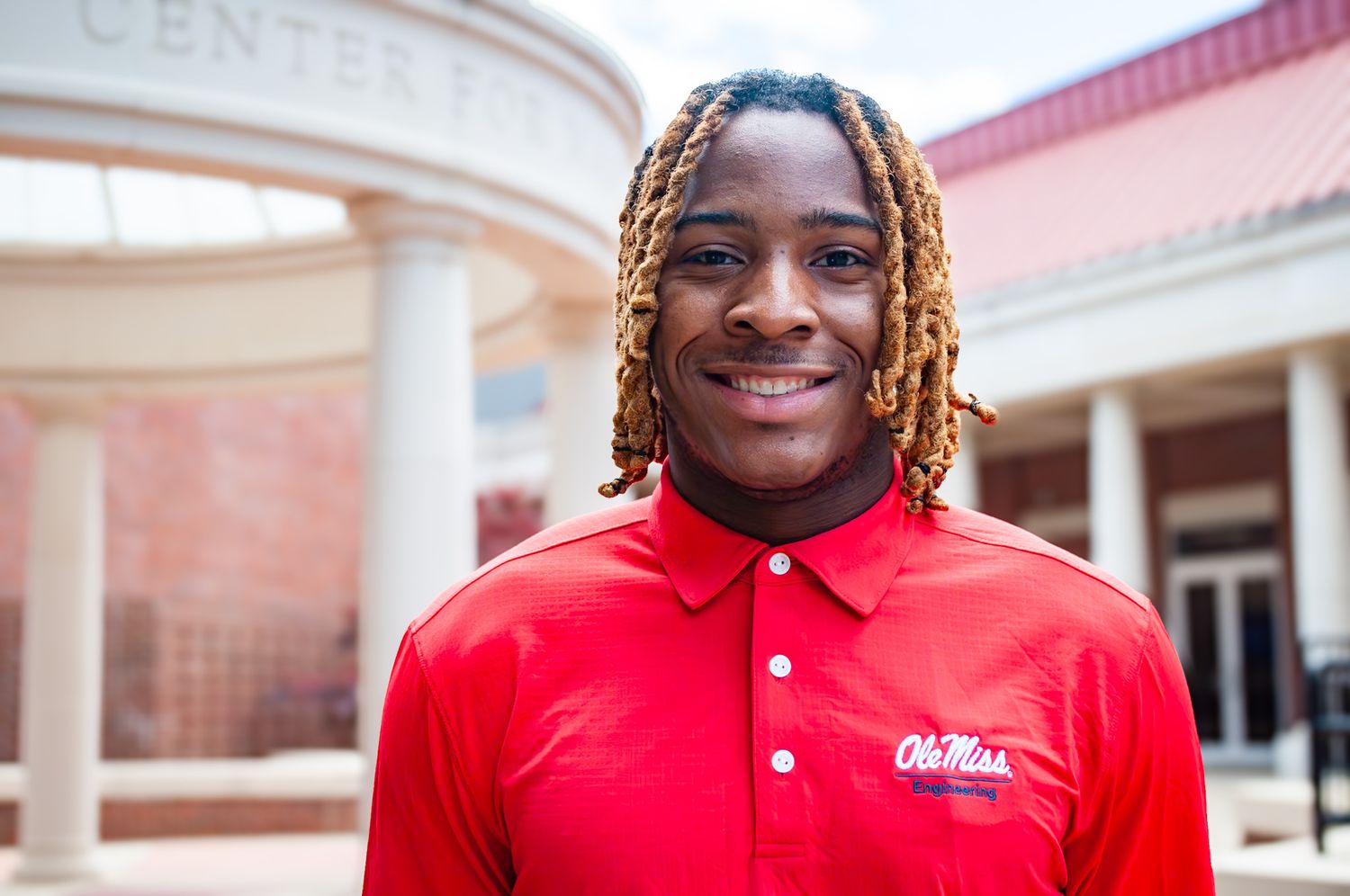 Young man in red Ole Miss polo shirt smiling outdoors near a campus building on a sunny day.