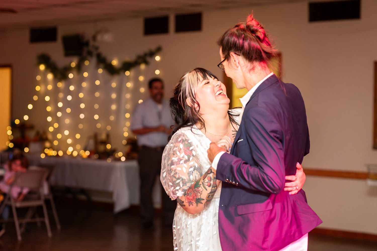 Couples slow dance together on the dancefloor at an evening reception with warm lighting and twinkle lights in background.