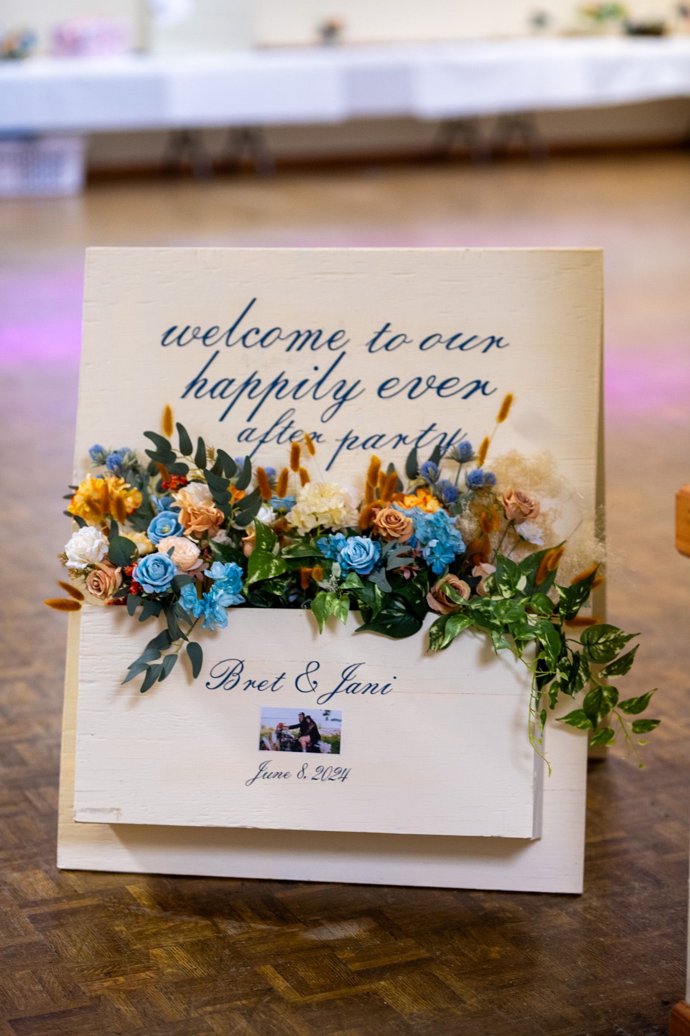 Wedding welcome sign decorated with blue and orange flowers and greenery displays 'Welcome to our happily ever after' text.