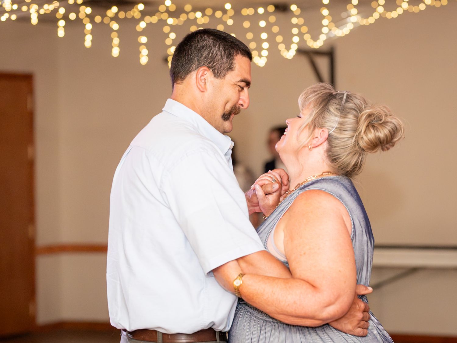 A couple dancing close together under warm string lights at an indoor reception venue.