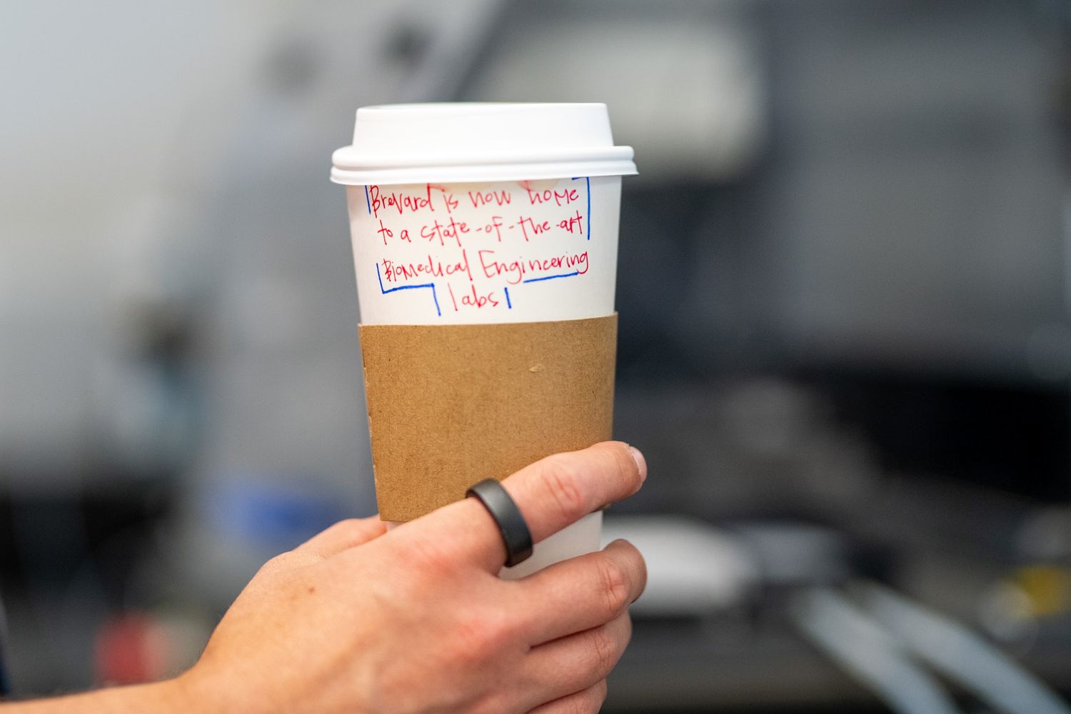 Hand holding a white disposable coffee cup with cardboard sleeve and handwritten notes in red marker.