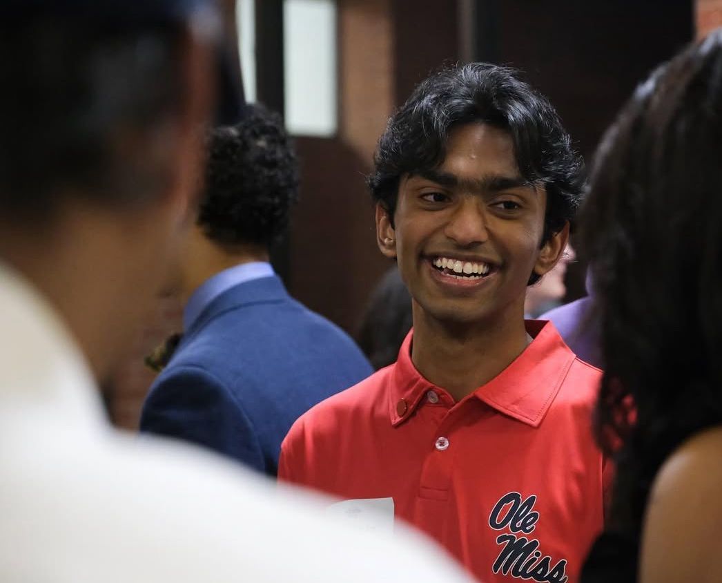 Smiling young man in red Ole Miss polo shirt socializing at an indoor gathering or event.