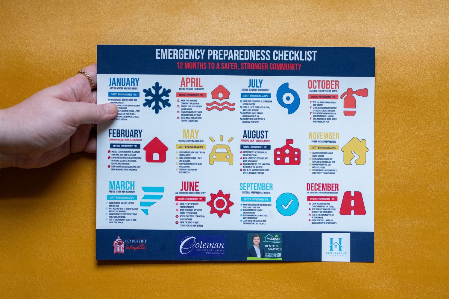 Hand holding an Emergency Preparedness Checklist card with monthly safety tips and colorful icons for disaster readiness.