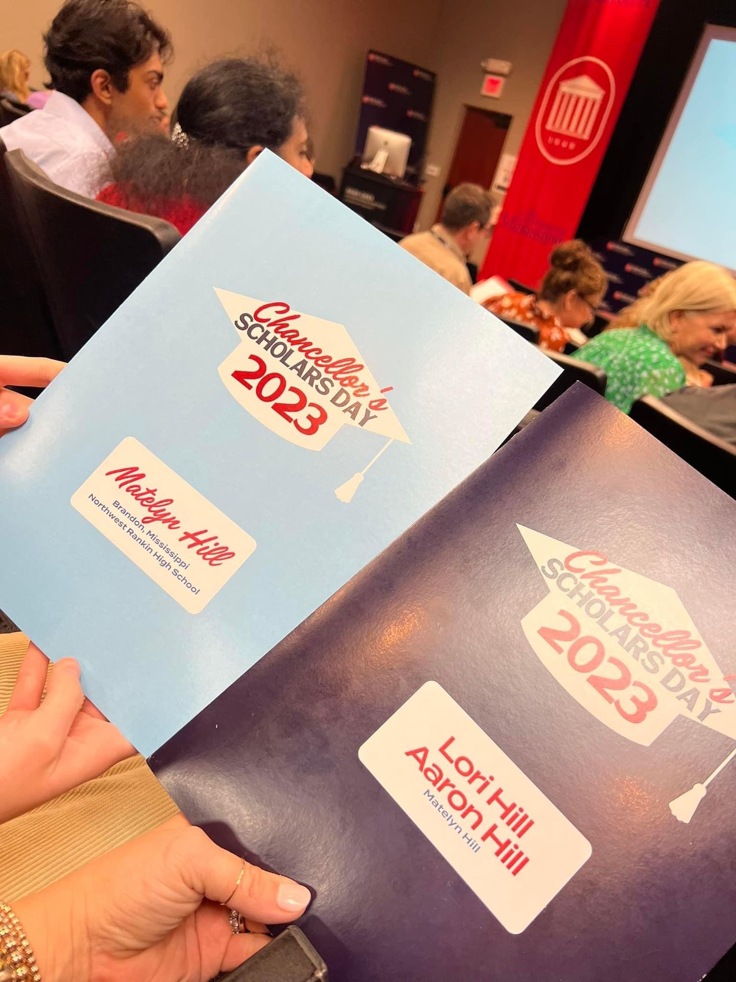 Two Chancellor's Scholars Day 2023 folders held up, one labeled Madelyn Hill and another Lori Hill and Aaron Hill.