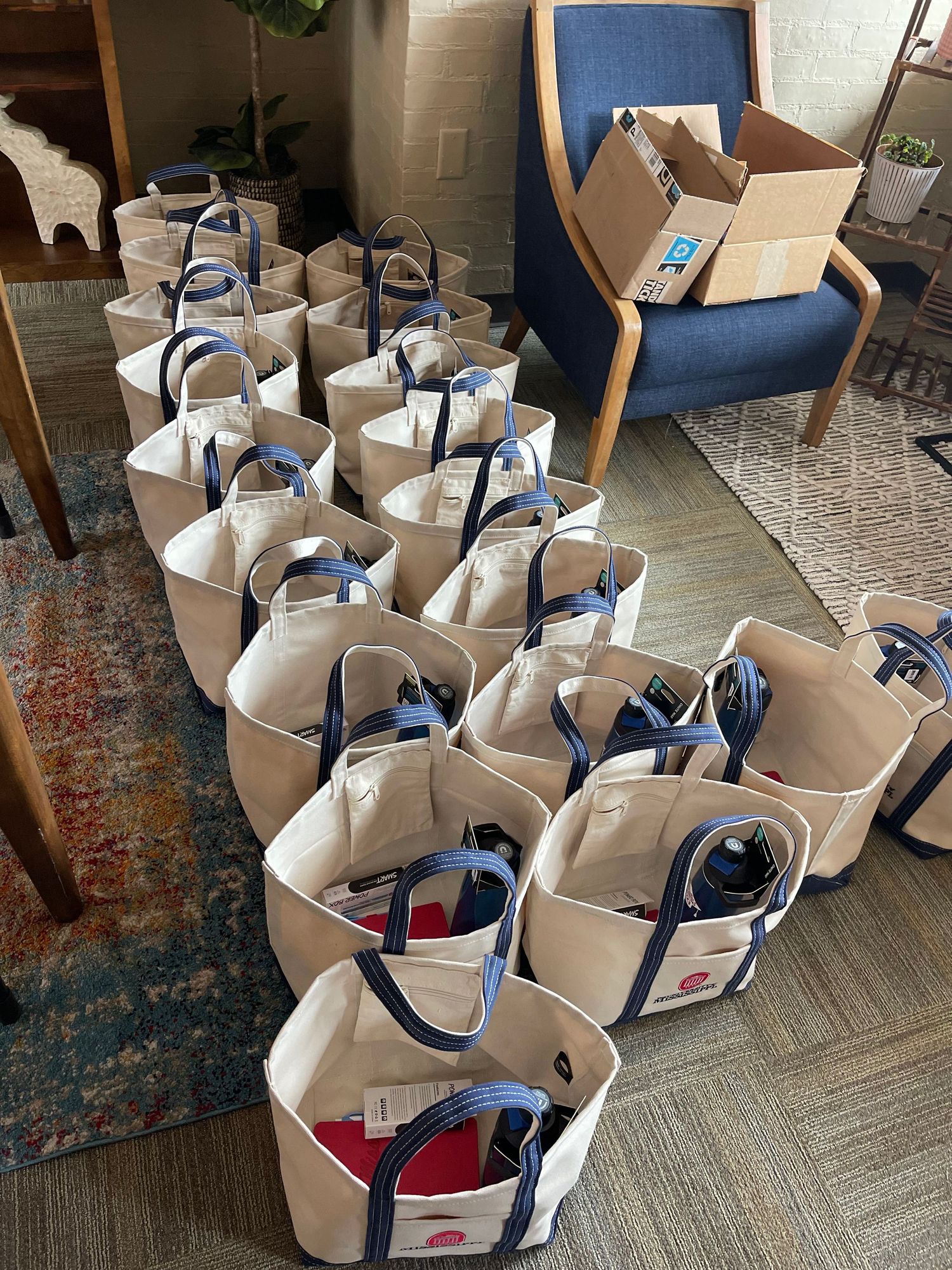Multiple canvas tote bags with blue handles arranged on a floor, filled with items, next to a blue chair and cardboard box.