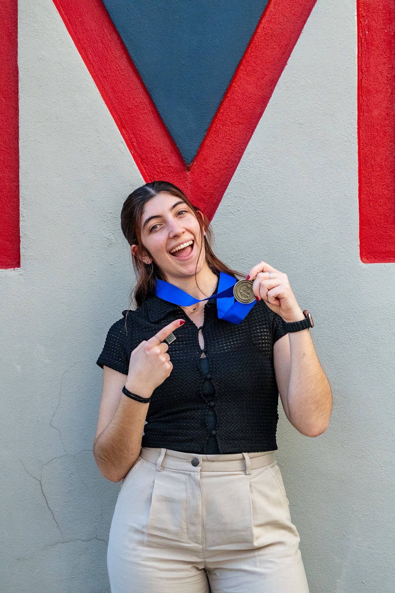 Young woman smiling proudly while pointing at a gold medal with blue ribbon, standing against a colorful wall.