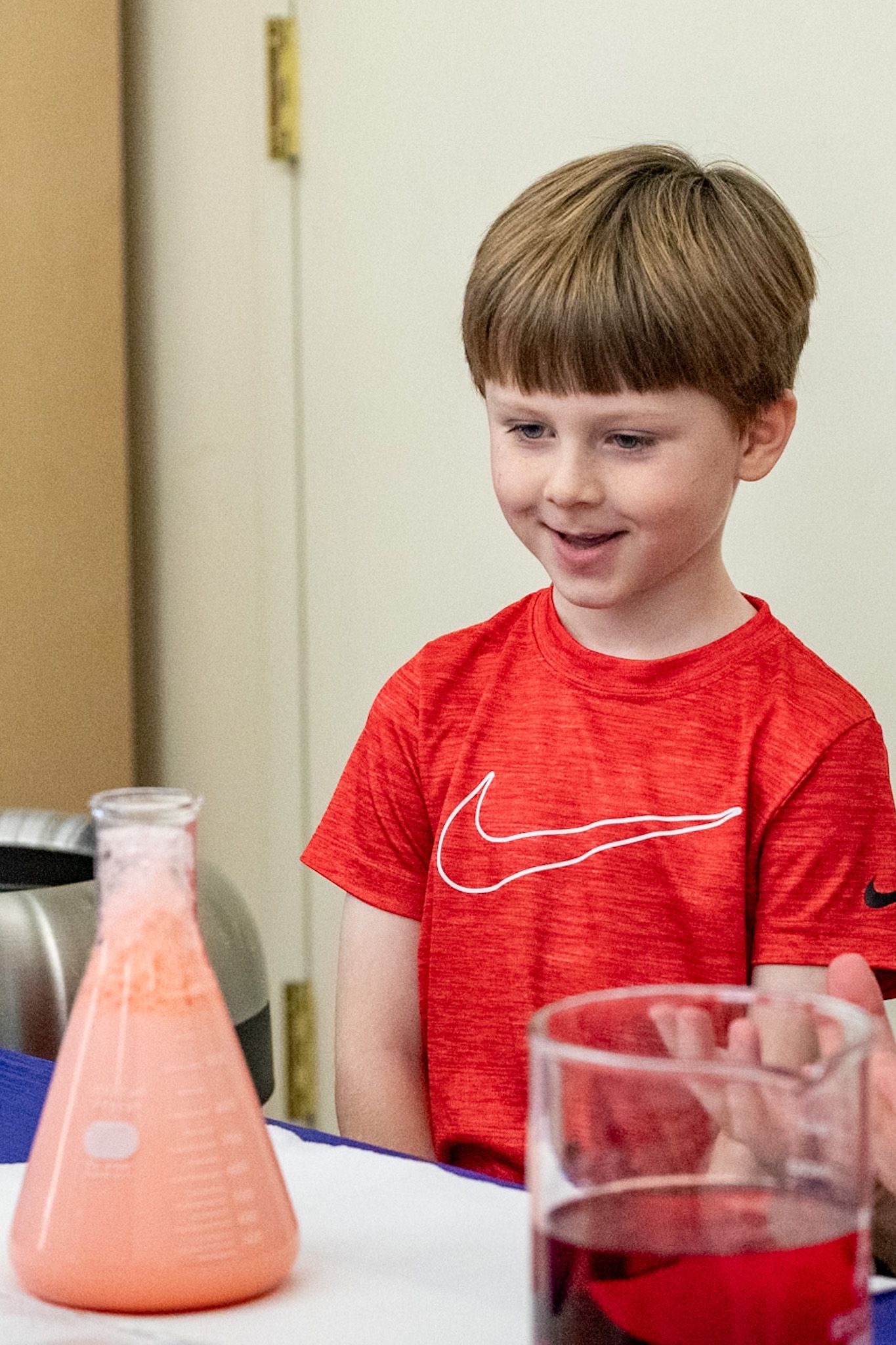 Young boy in red Nike shirt smiling at a pink liquid in an Erlenmeyer flask during a science experiment.