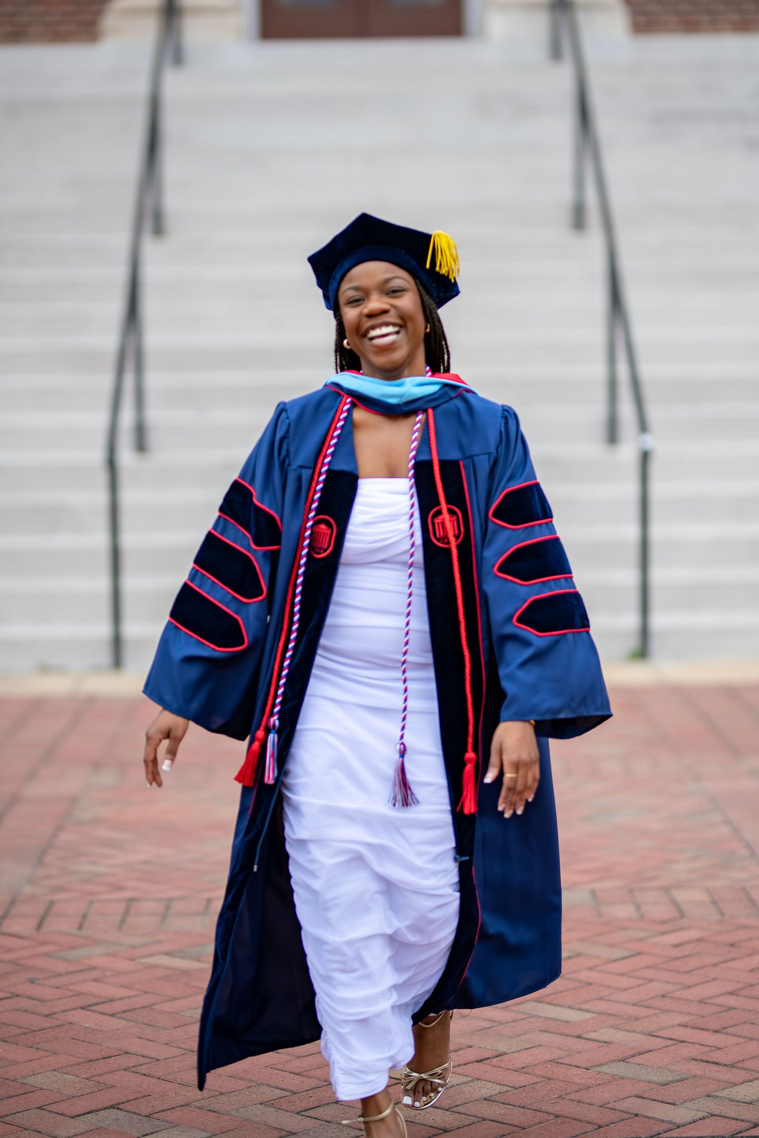 Graduate in blue academic regalia stands on brick path against building steps for celebratory photographs.