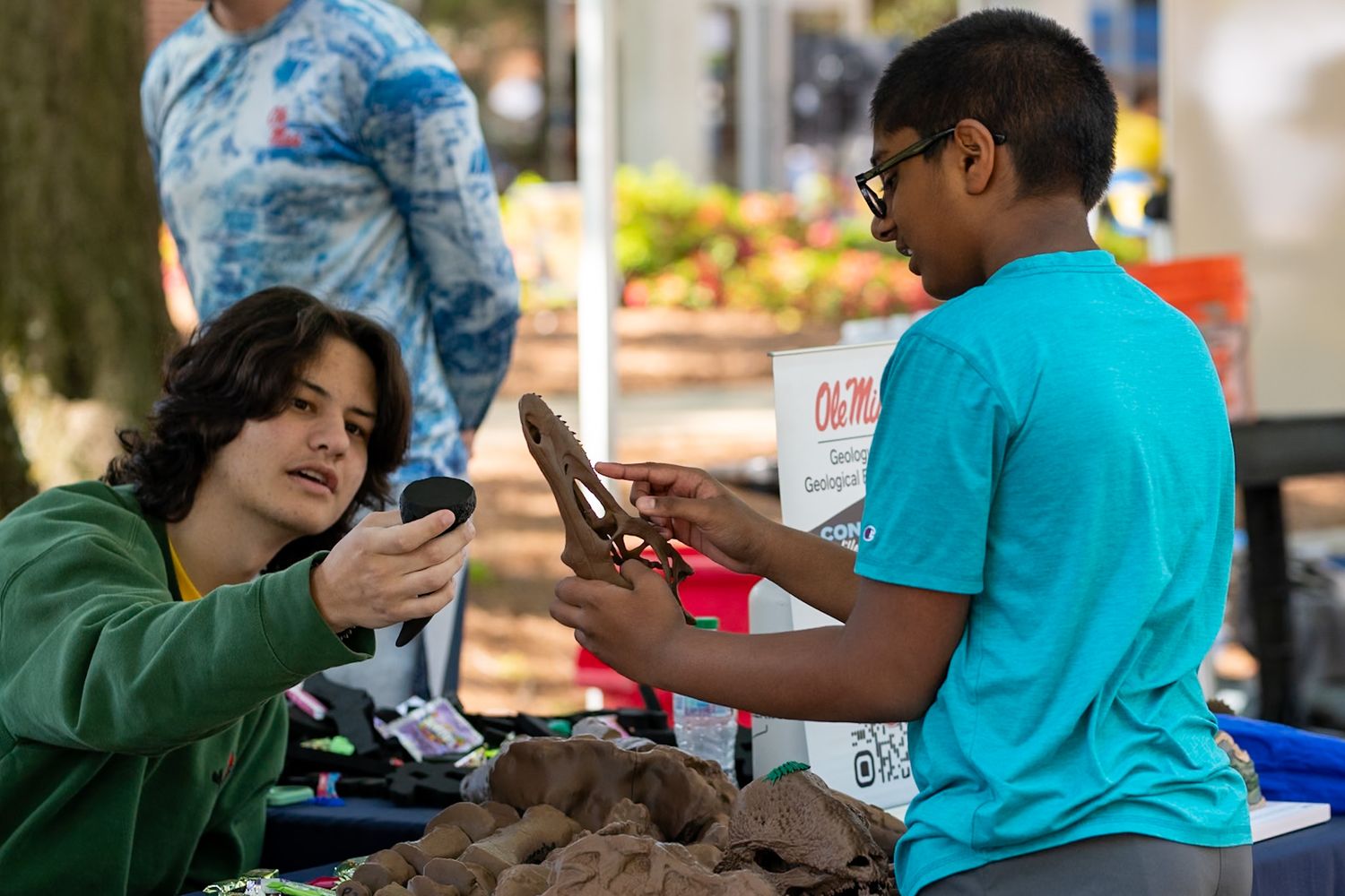 Two kids examining produce at an outdoor farmers market booth with vegetables displayed on the table.