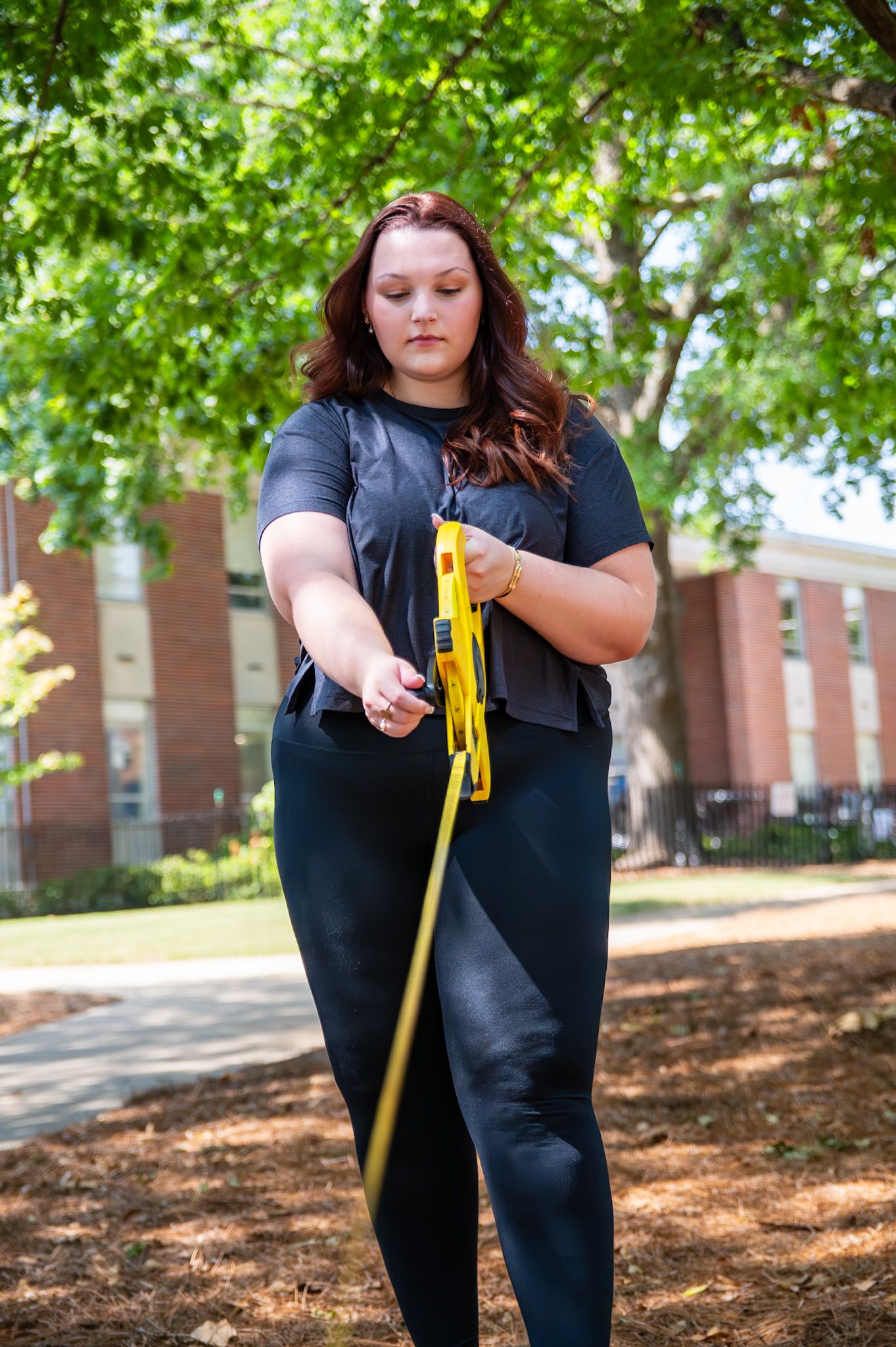 Woman in black athletic wear uses yellow resistance band outdoors near brick building.
