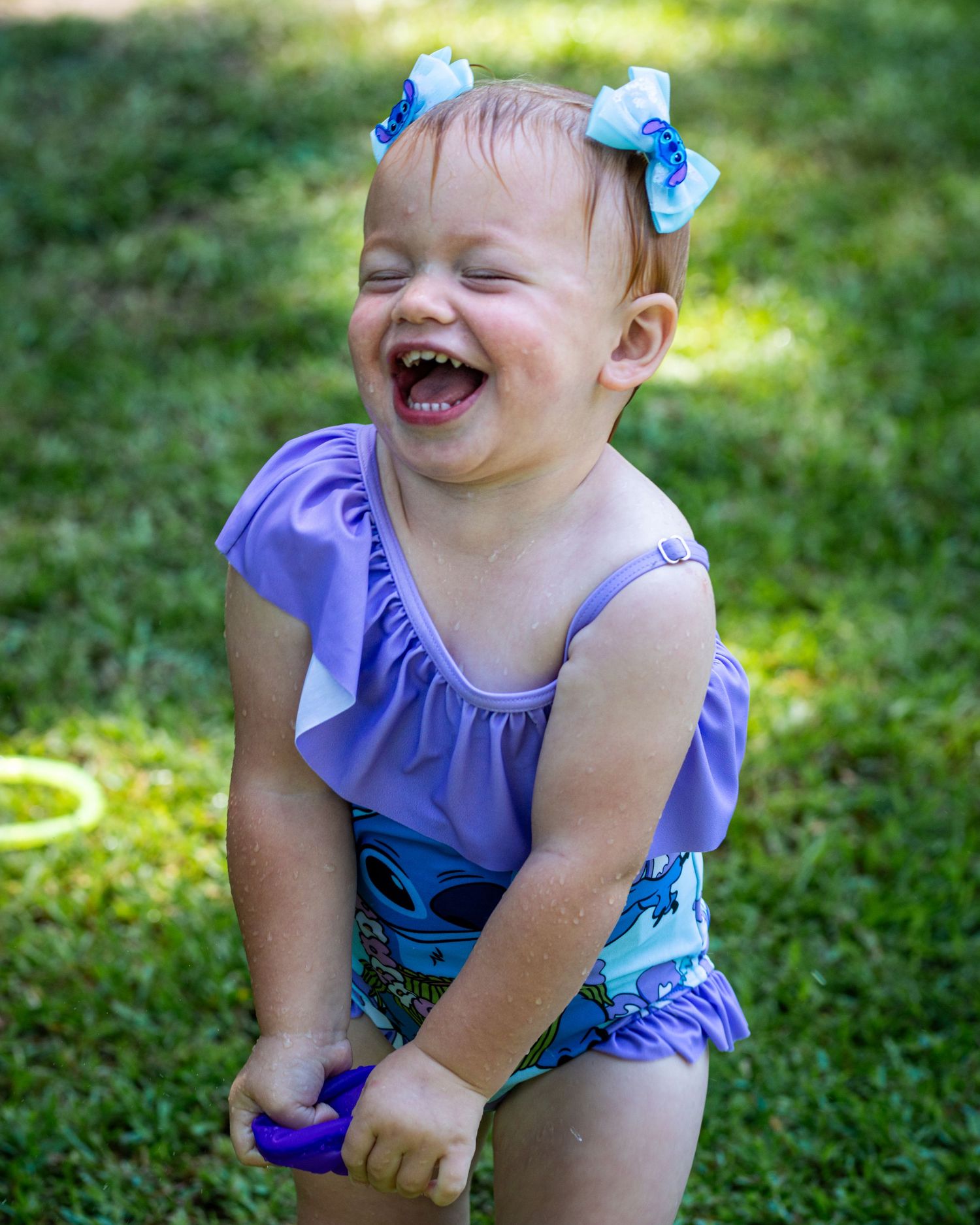 Laughing baby girl in purple Stitch swimsuit and blue hair bows plays outdoors on green grass.