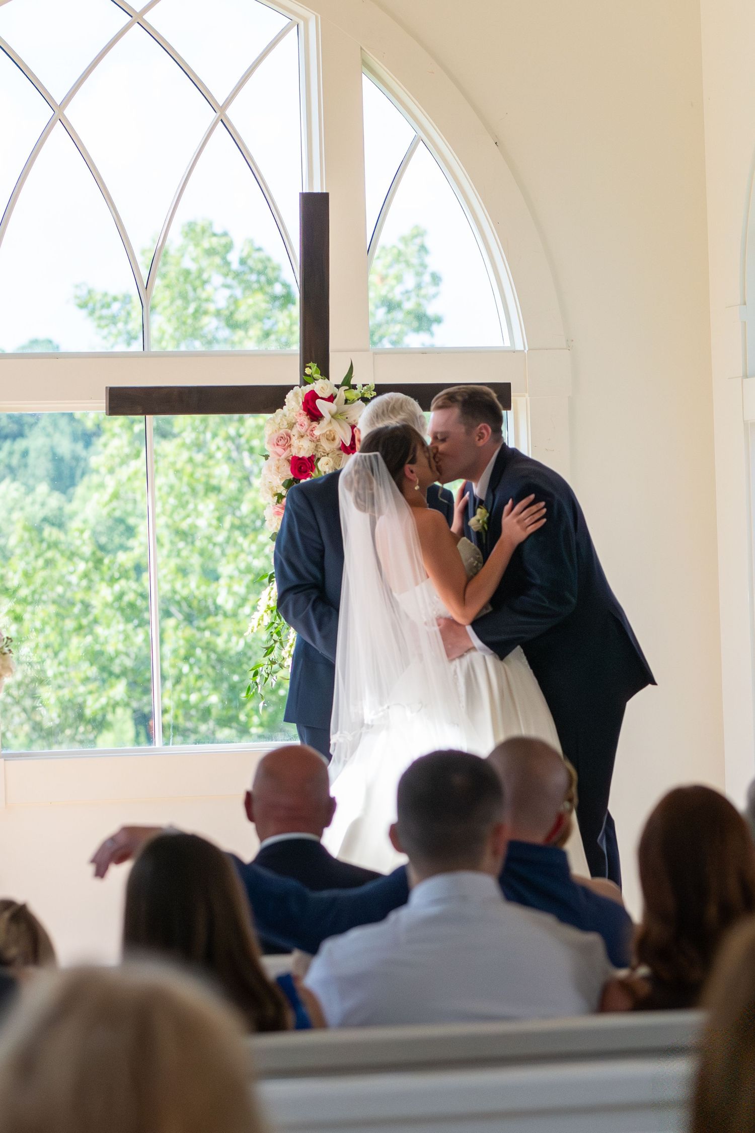 Bride and groom share first kiss at chapel altar with floral bouquet and wooden cross before wedding guests.