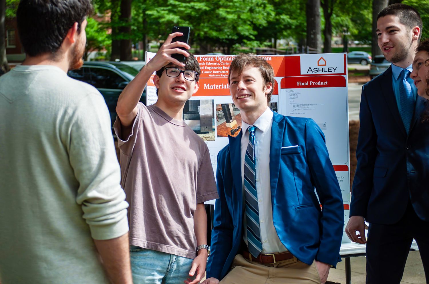 Students taking a selfie outdoors near an Ashley project display board at a college engineering or design expo.